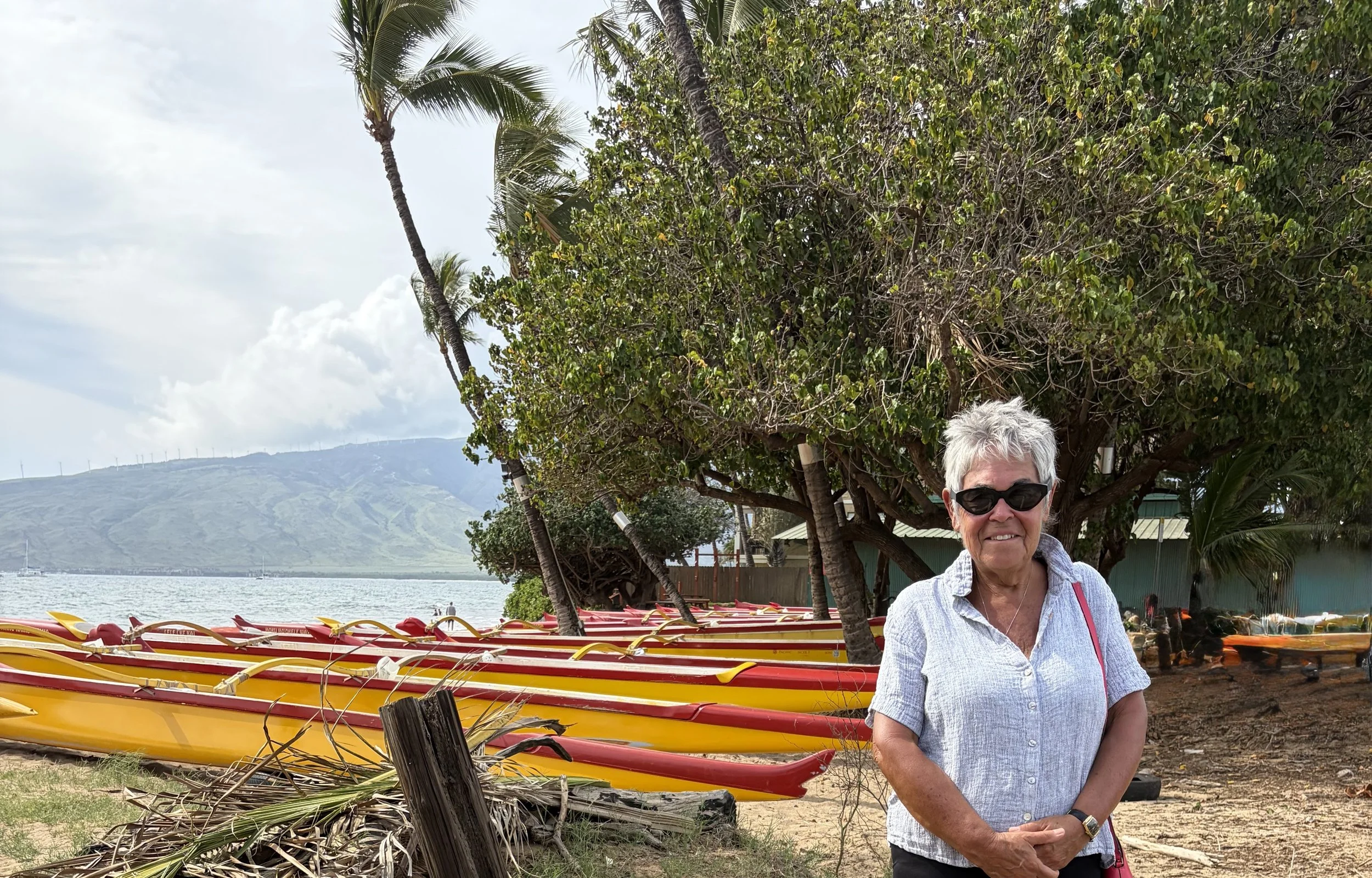 A woman with short hair wearing sunglasses and a light-coloured collared shirt, standing outdoors near a row of yellow and red kayaks, with lush green trees and a waterfront with sailboats in the background.