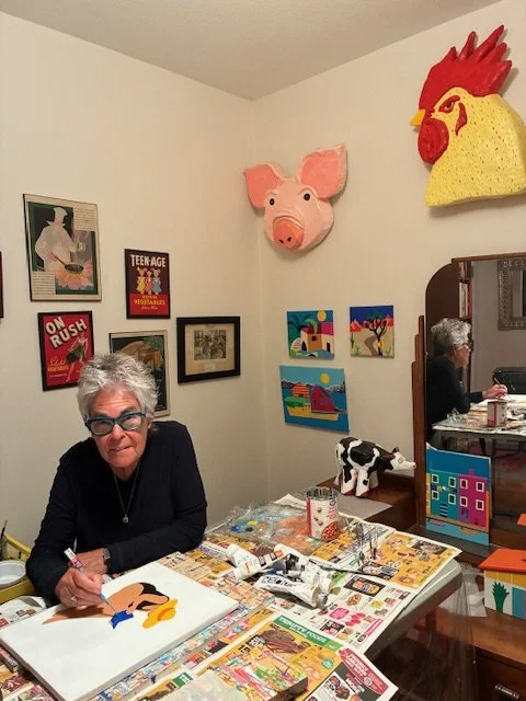 A woman sitting at a table painting a colorful abstract artwork, surrounded by art supplies and newspapers, with decorated walls featuring art.