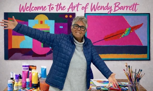 Woman with short hair and glasses smiling, standing with arms open in front of colourful artwork and art supplies, in an art studio or gallery.