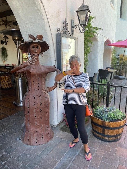 A woman standing next to a metal sculpture of a witch with a hat, on a sidewalk outside a building