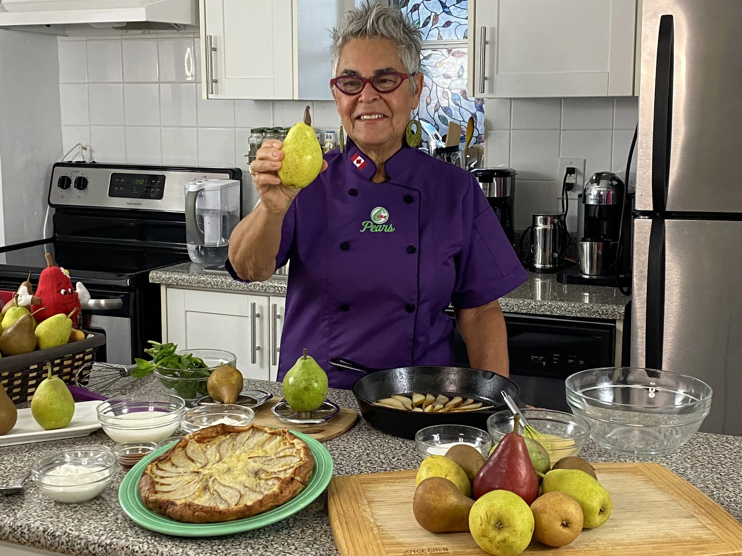 A woman with short hair, glasses, and a purple chef's coat holding a pear in a kitchen. The counter is filled with various fruits, bowls, and a pie, indicating food preparation or cooking.