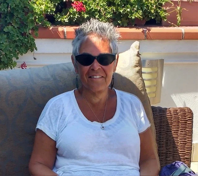 Woman with short hair, wearing sunglasses, a white t-shirt, sitting outdoors on a cushioned chair, with plants and flowers in the background.