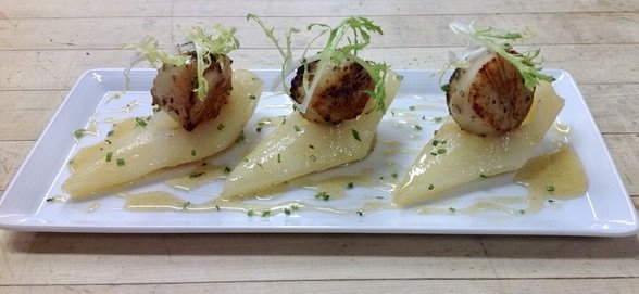 Three pieces of seared scallops on slices of potatoes, garnished with lettuce and herbs, served on a white rectangular plate.