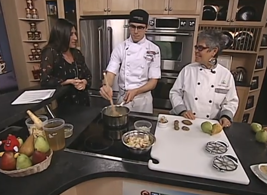 Three people in a kitchen, one in a chef's coat cooking on a stove, another woman watching, and a second chef in a white coat standing next to a cutting board with food ingredients.