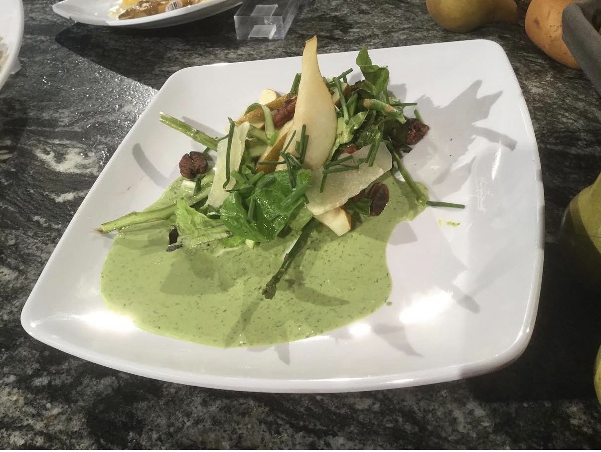 A plated salad with greens, sliced pears, candied walnuts, and green herb dressing on a white square plate.
