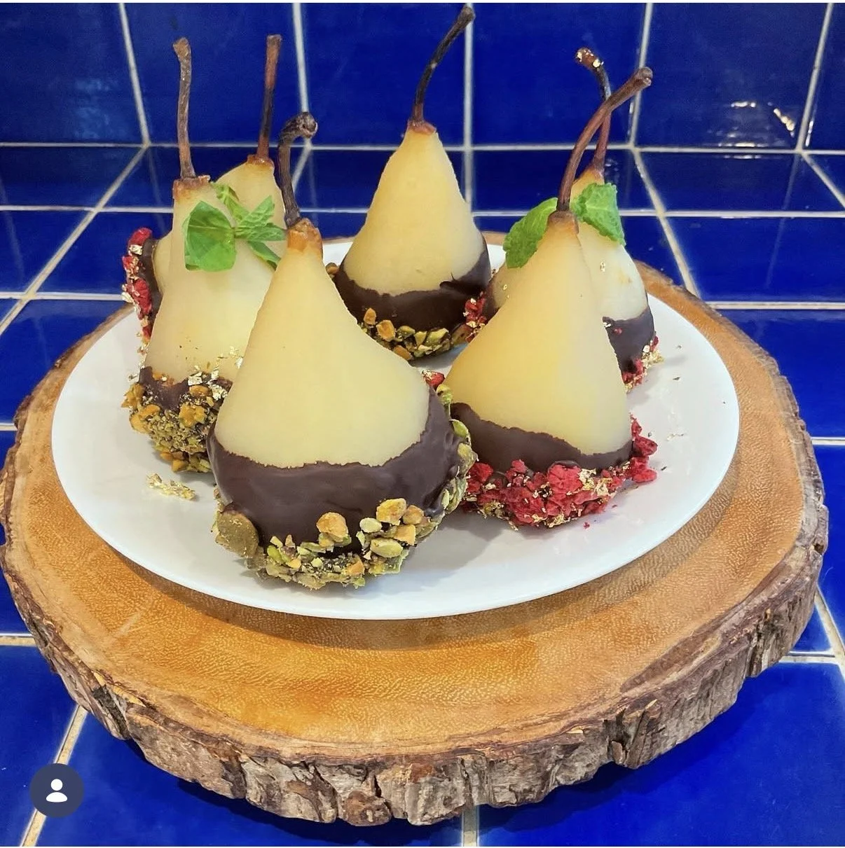 Five pear-shaped desserts coated in dark chocolate at the bottom, decorated with crushed nuts and dried berries, with small green mint leaves on top, arranged on a white plate on a wooden slab with a blue tiled background.