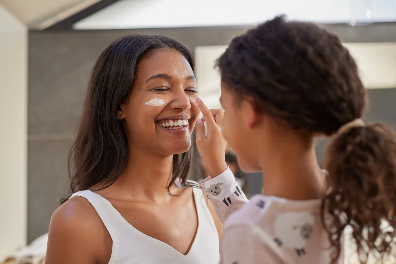 A young girl applies sunscreen to a smiling woman with long dark hair, indoors with natural light.