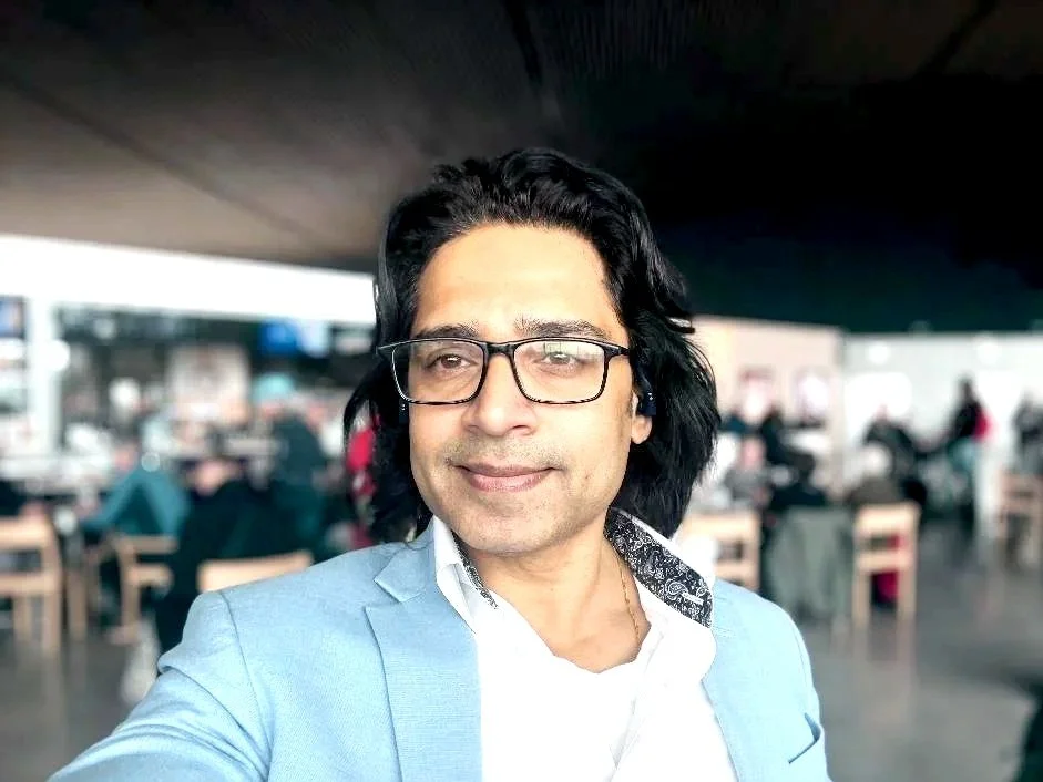 A man with black hair and glasses taking a selfie in a restaurant or cafe, with tables and people in the background.