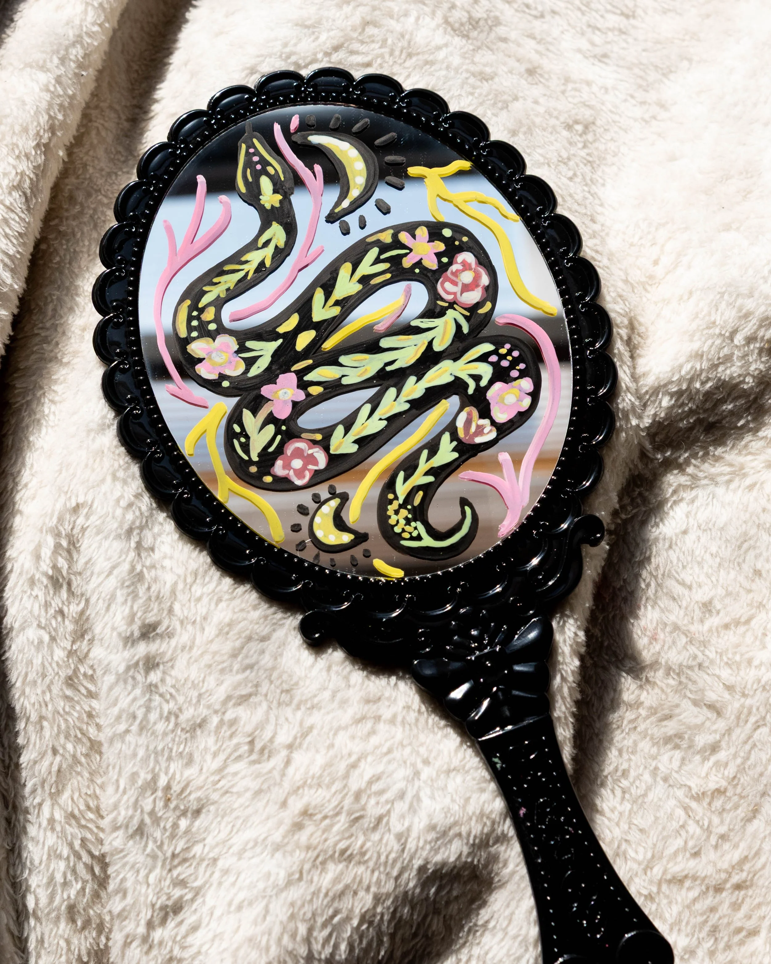 Decorative hand mirror with a black ornate frame, featuring a colorful painted snake design on the reflective surface, placed on a white fluffy fabric background.