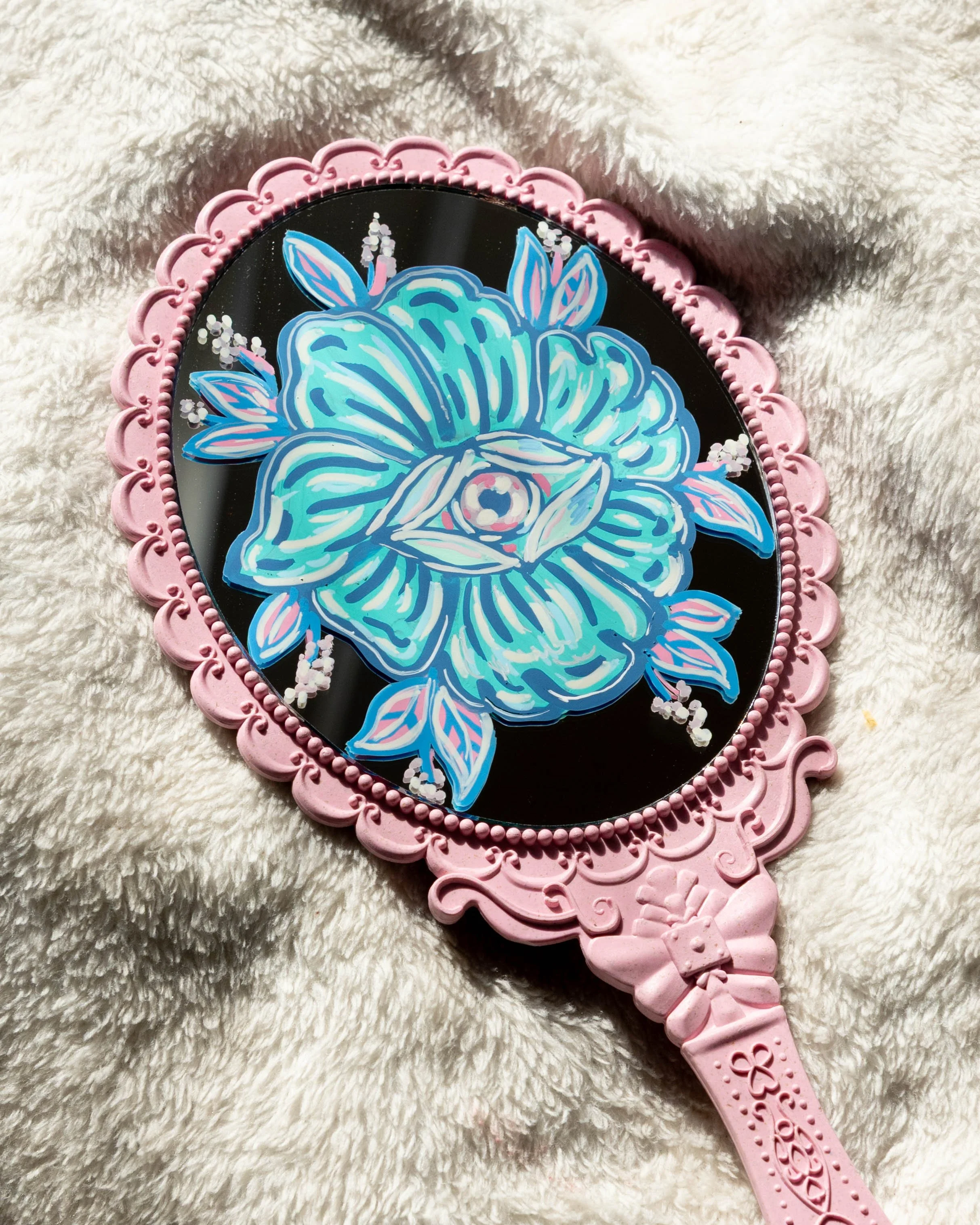 Pink hand mirror with ornate handle, reflecting a stylized blue rose design with leaves and small flowers on a black background, resting on a soft white fabric.