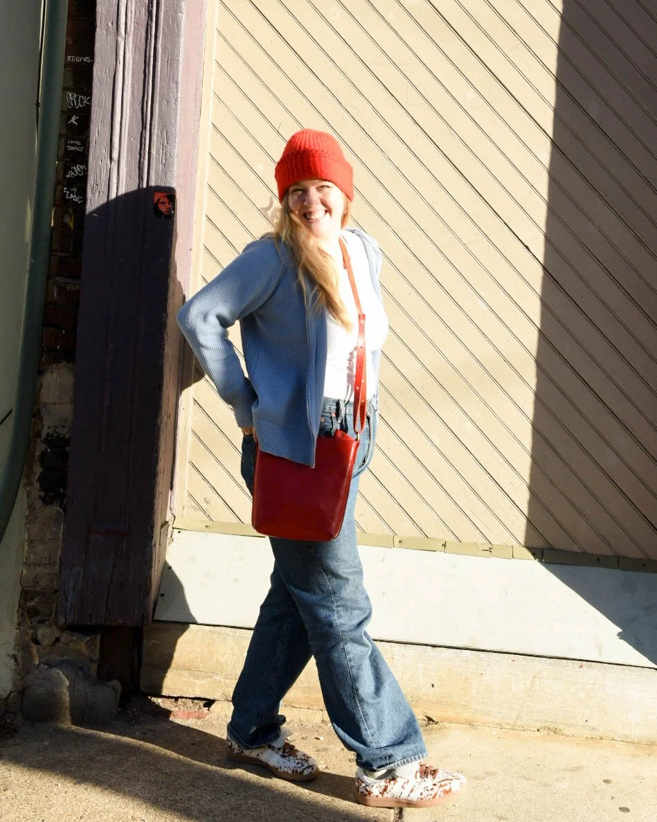 A woman smiling, wearing a red beanie, blue jacket, jeans, and sneakers, standing against a wall in sunlight.