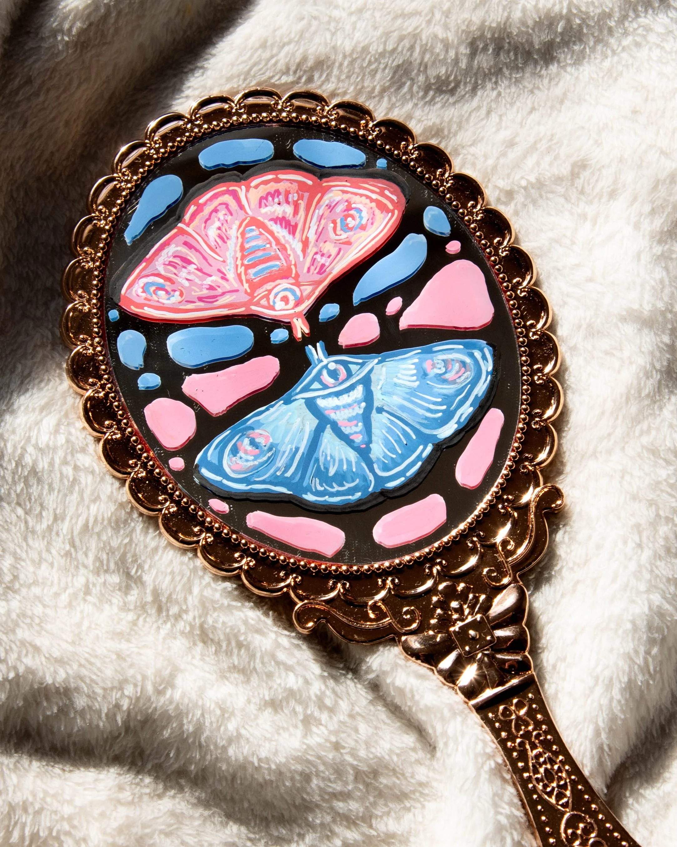 Decorative mirror with stained glass butterflies in pink and blue, surrounded by a rose gold frame, resting on a soft, cream-colored fabric.