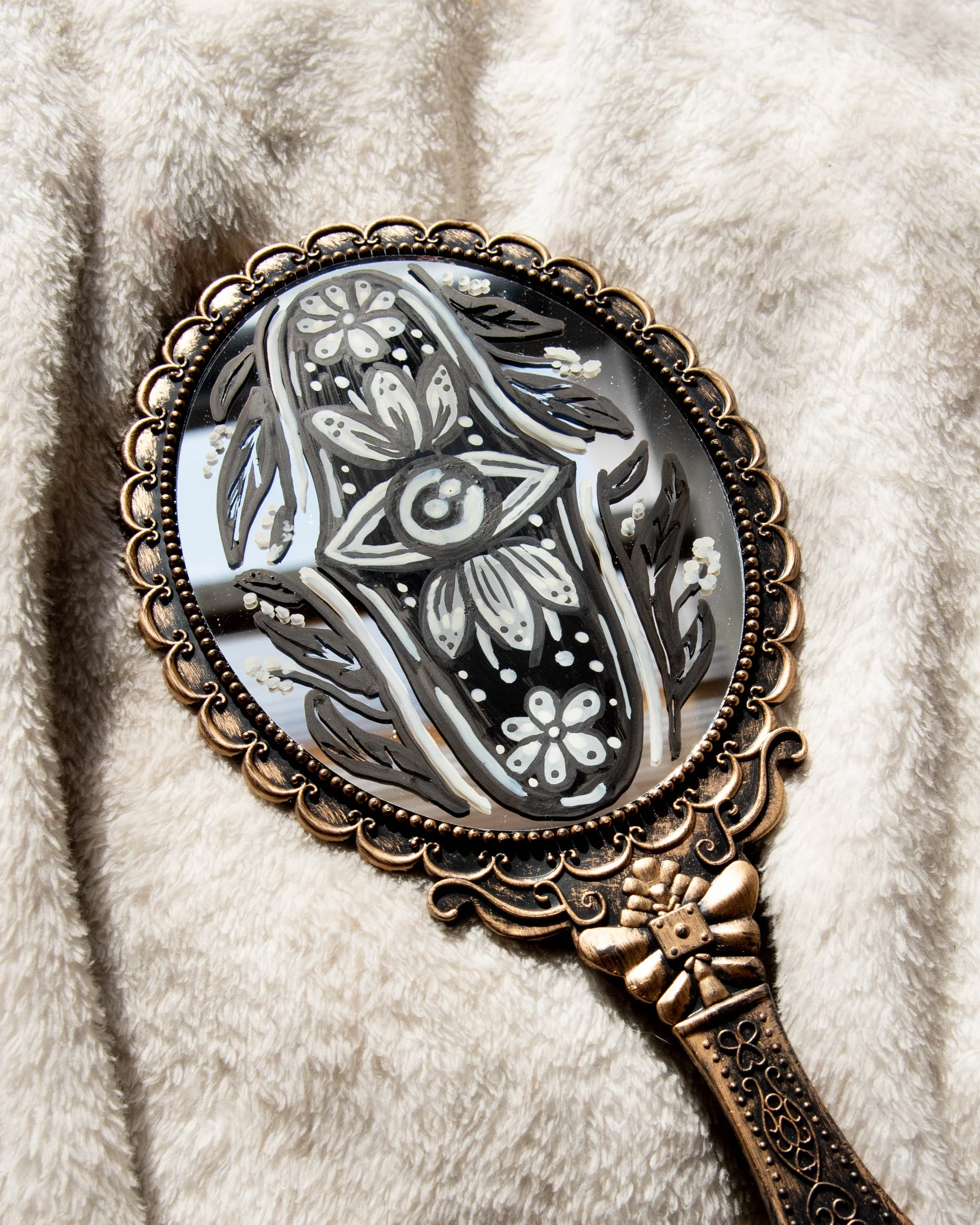 Decorative hand mirror with floral and geometric patterns in black, white, and bronze color, resting on a light-colored, fluffy fabric.