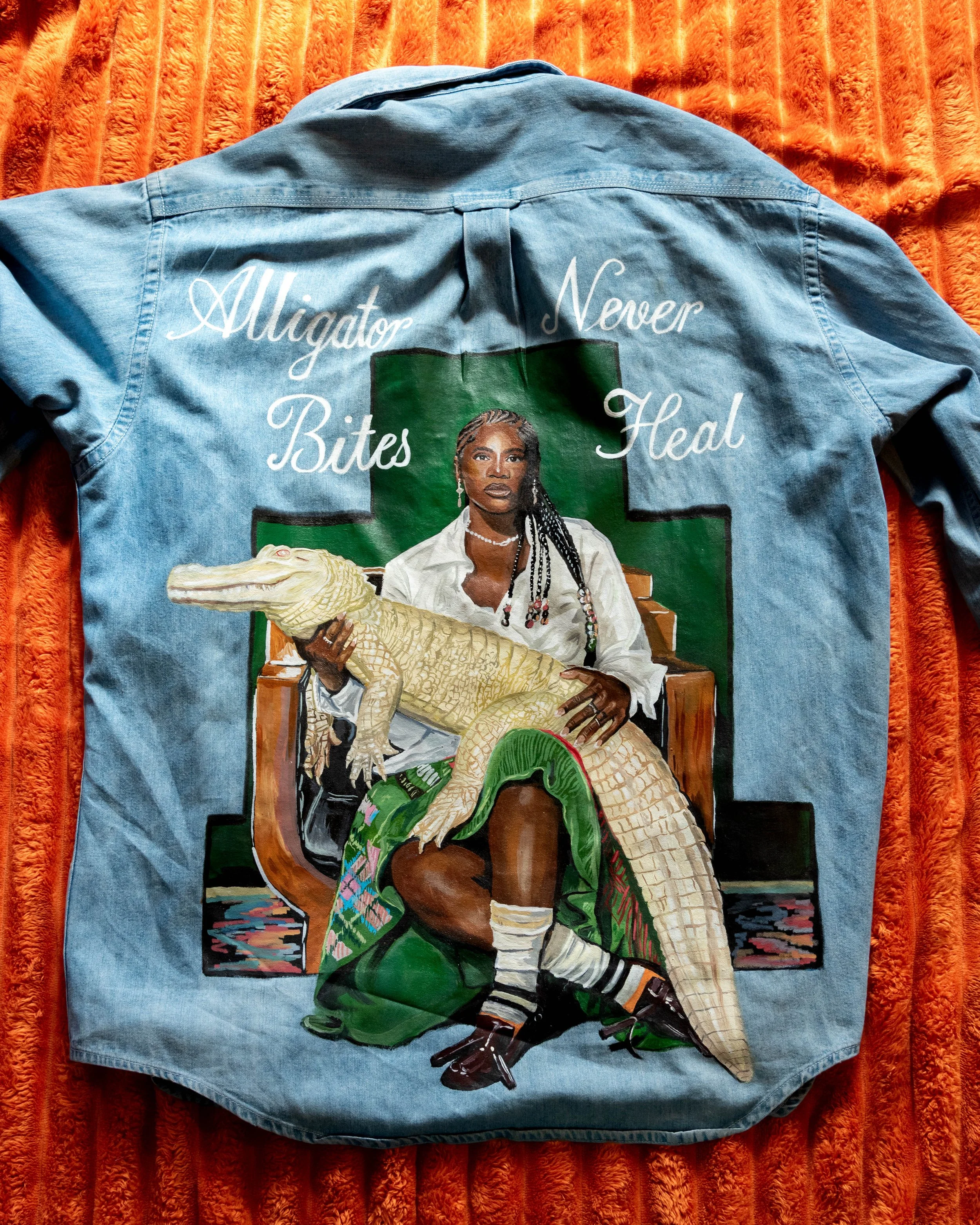 A denim jacket with a painted portrait of a woman with braids, sitting on a chair, holding an alligator. The jacket has the phrases "Alligator Never Bites" and "Heal" written on it, with a green cross in the background, all on an orange textured fabric.