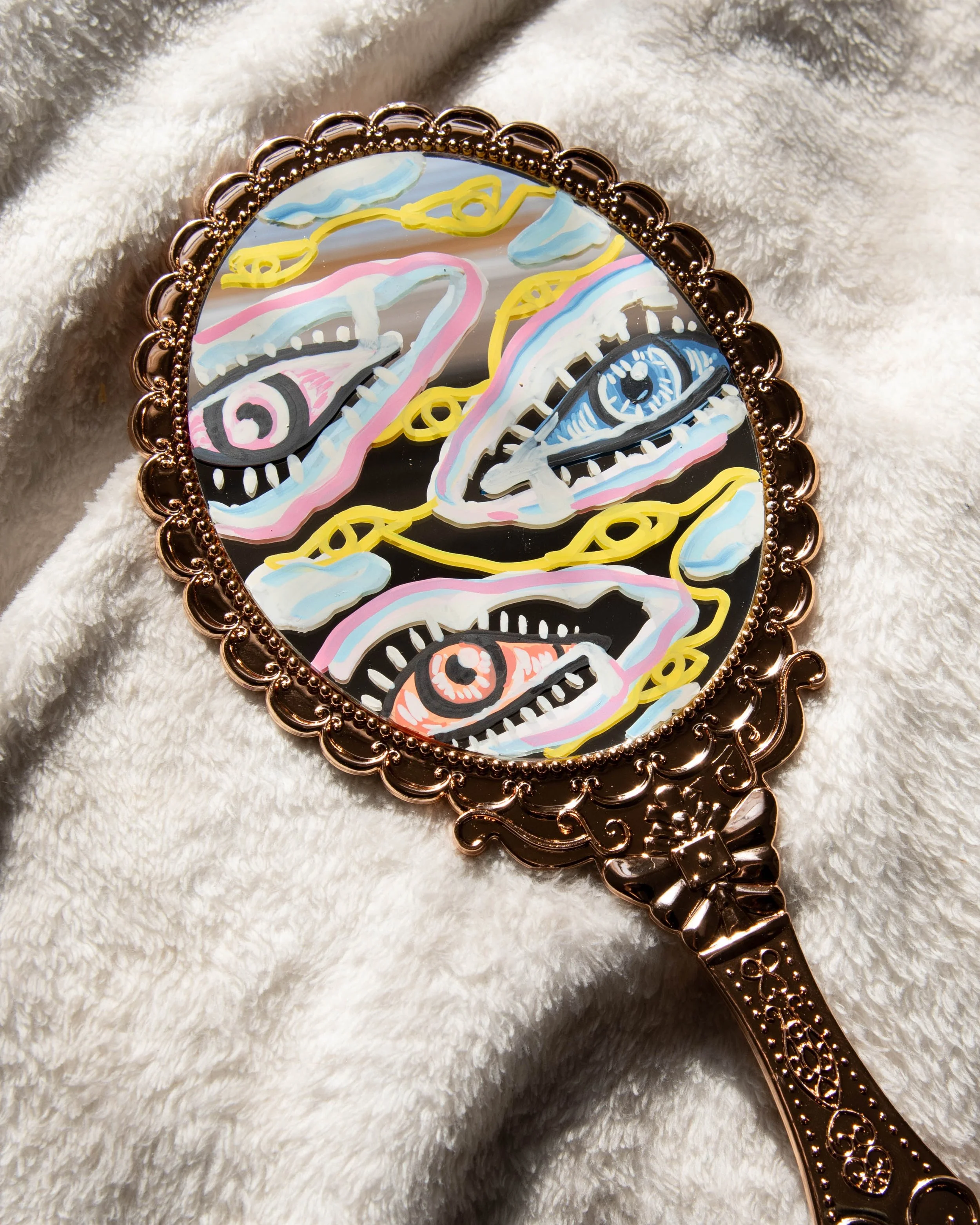 Decorative mirror with abstract designs of eyes and swirling patterns in pastel colors, gold accents, and intricate border on a soft, cream-colored fabric background.