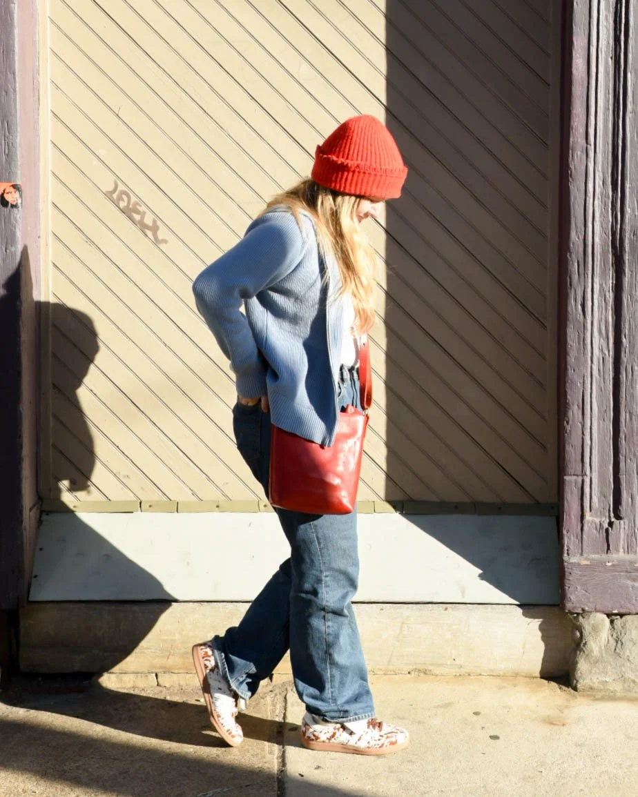 A woman wearing a red knit hat, blue jacket, jeans, and patterned sneakers walking past a wooden building with a beige door.