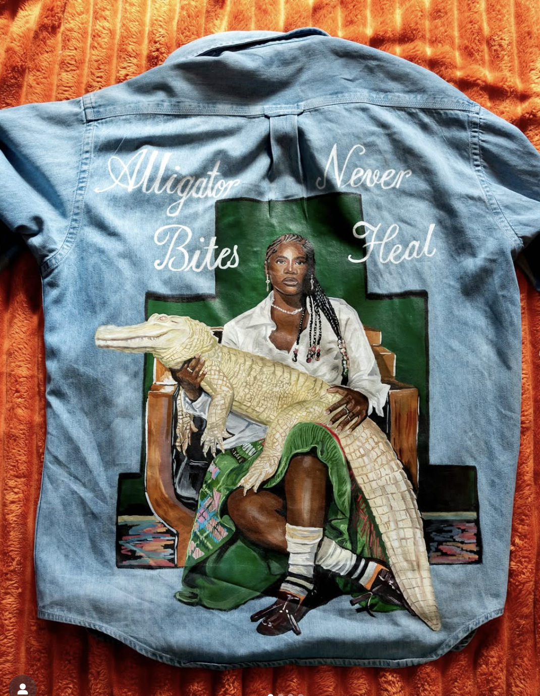 A painted denim jacket featuring a woman with braids, sitting on a green background with a crocodile in her lap. The jacket has the words 'Alligator Never Bites Heal' written in white script.