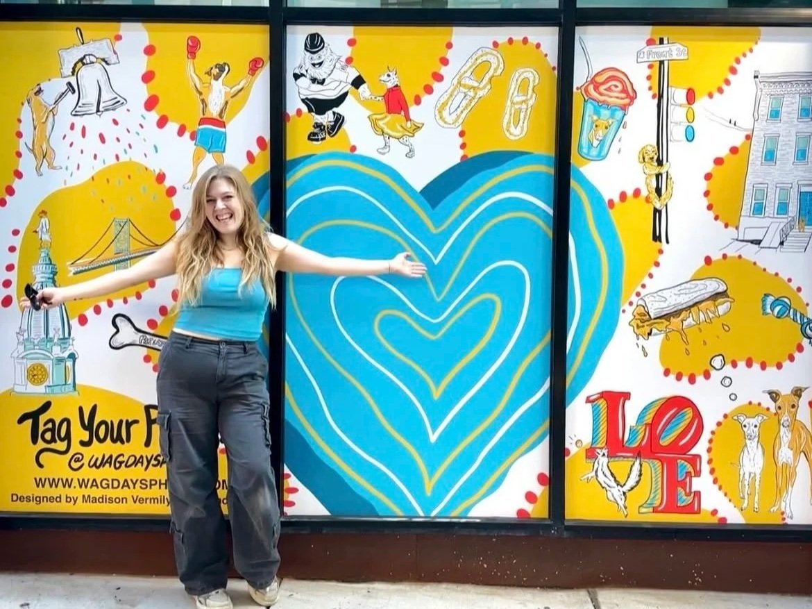 Young woman with long blonde hair smiling and extending her arms, standing in front of a colorful mural featuring hearts, a bridge, and various playful illustrations.