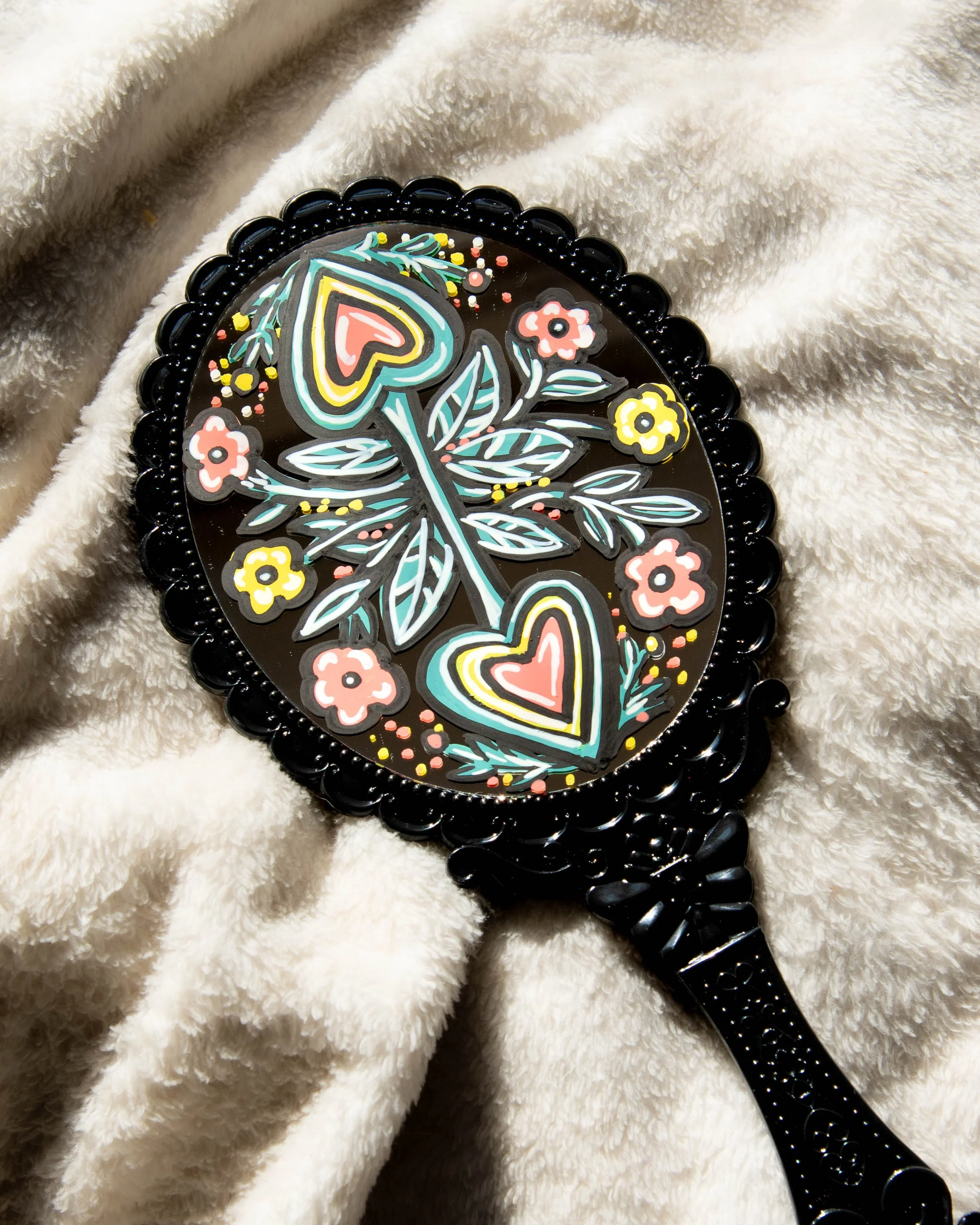 Decorative hand mirror with colorful floral and heart patterns on the reflective surface, resting on a soft cream-colored blanket.