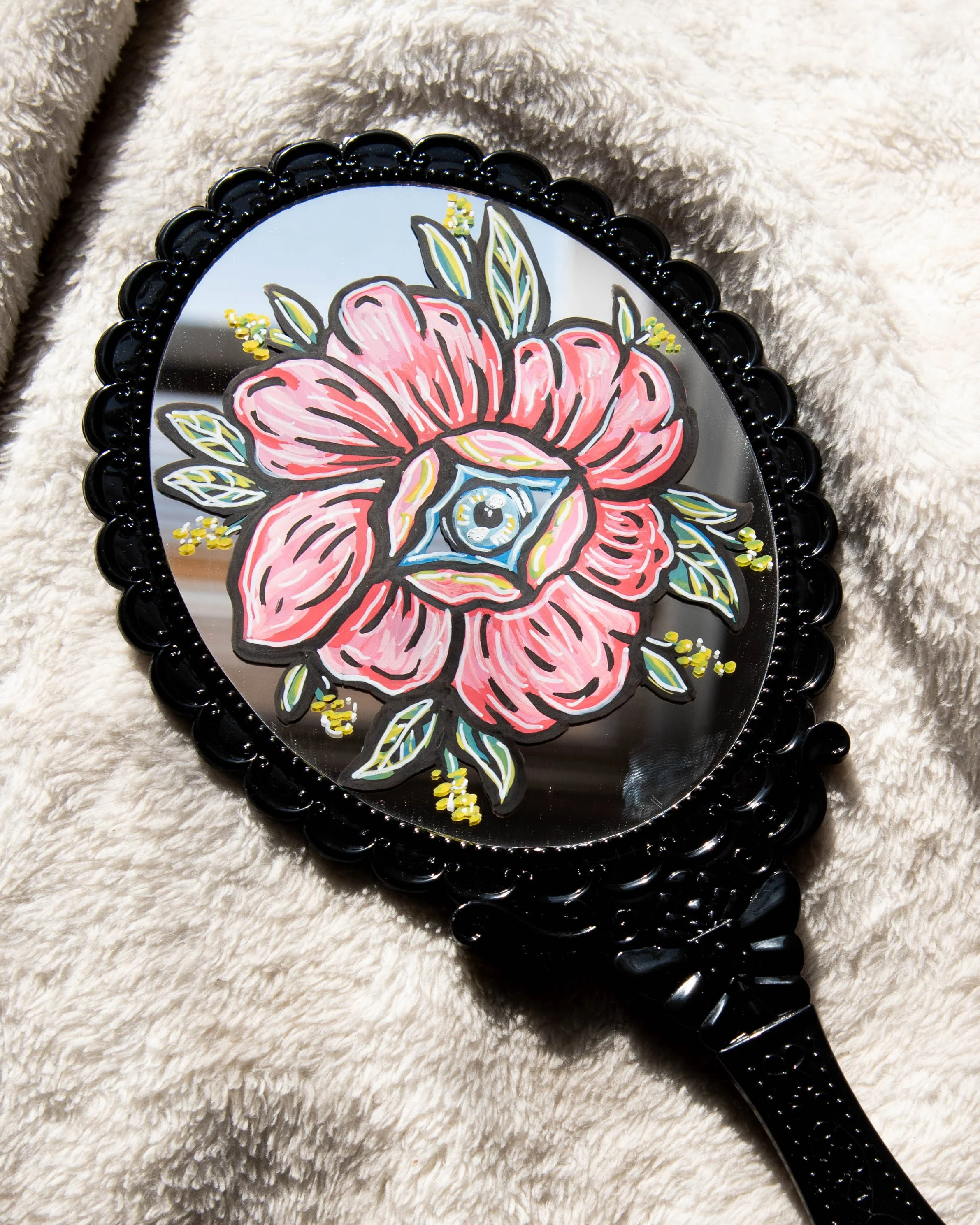 Decorative black handheld mirror with a painted pink flower and green leaves design on the reflective surface, placed on a beige, fuzzy fabric background.