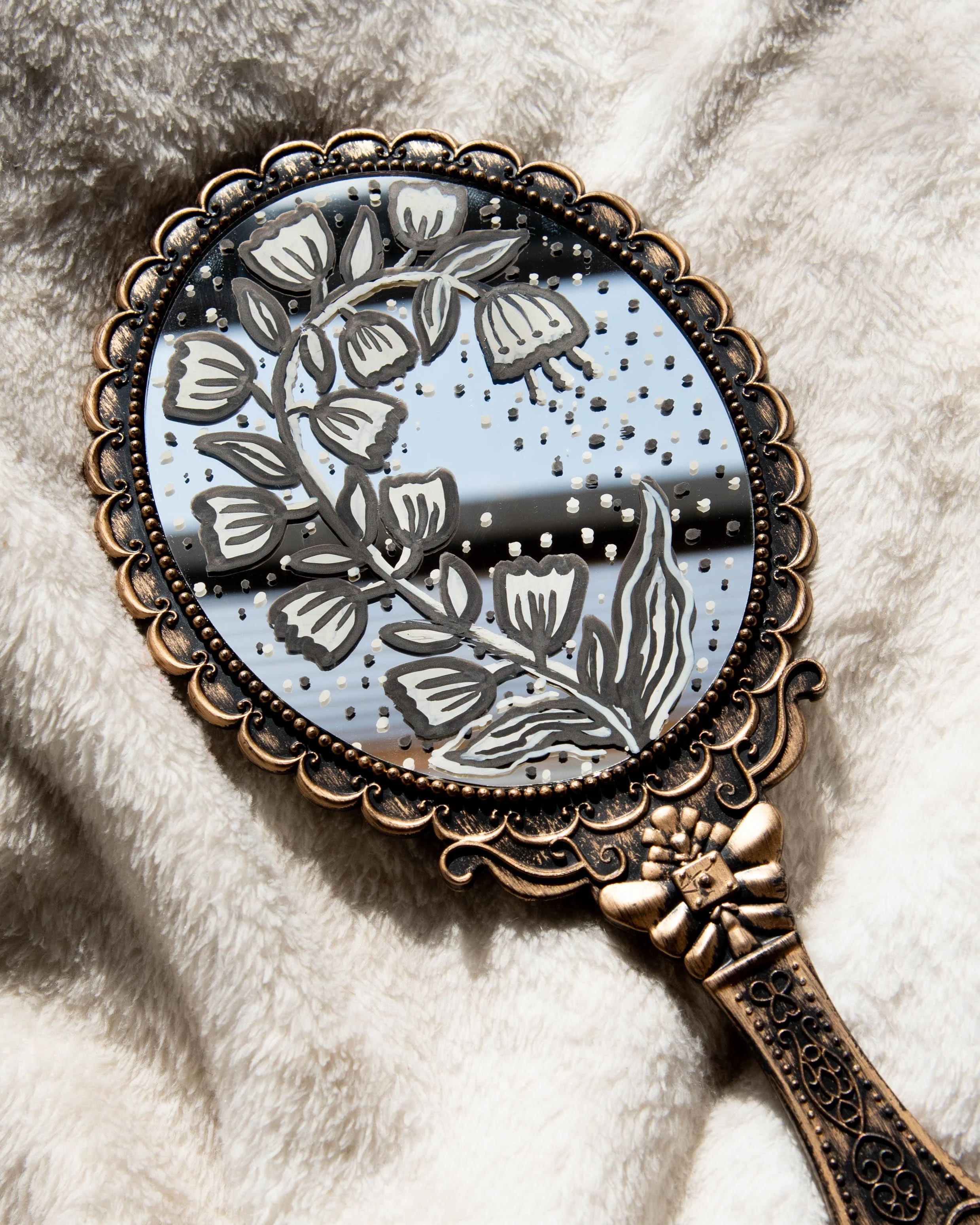 Decorative hand mirror with a floral design, lying on a soft beige fabric.