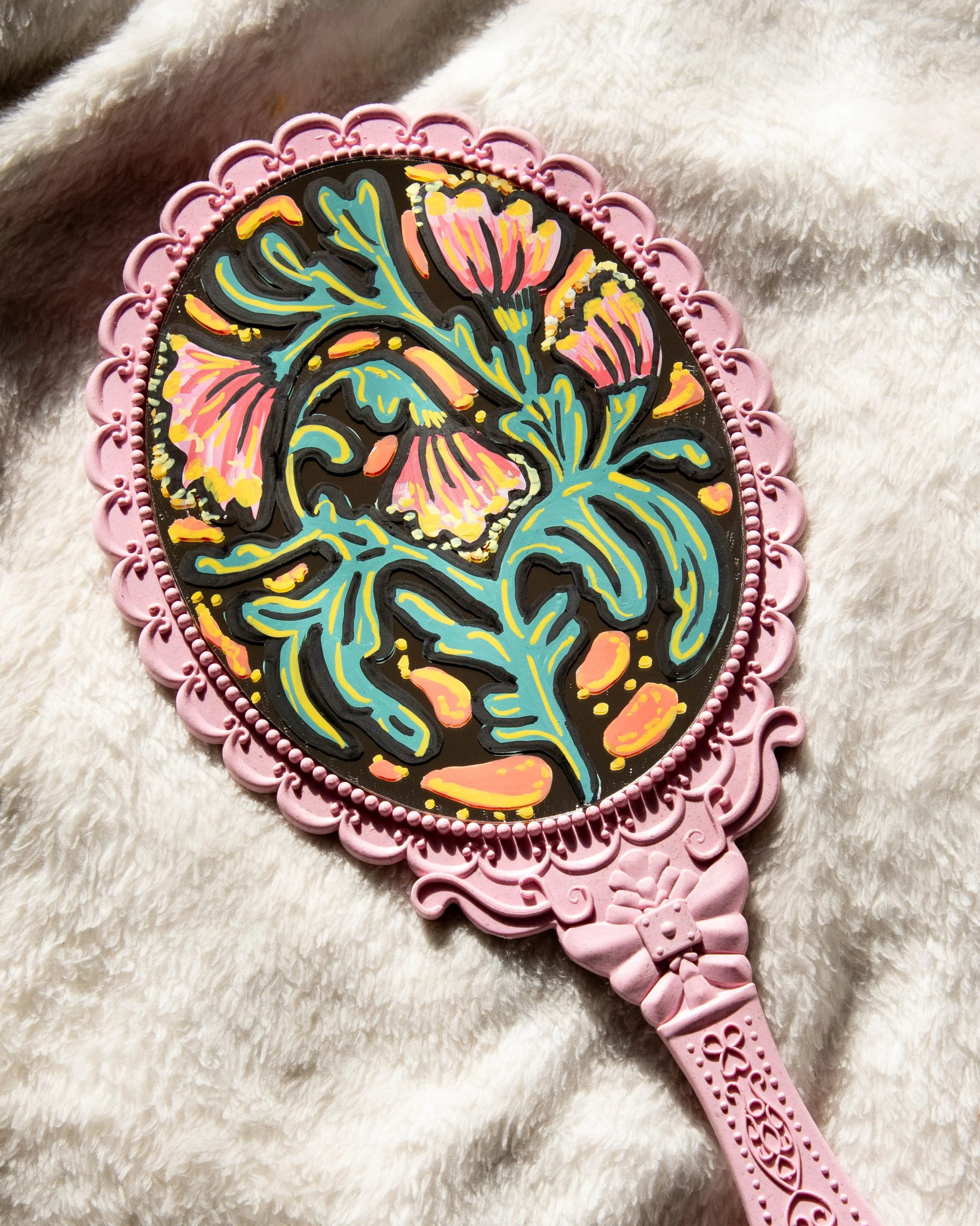 Pink hand mirror with decorative border on a soft, white fabric, featuring a colorful floral design on the mirror surface.