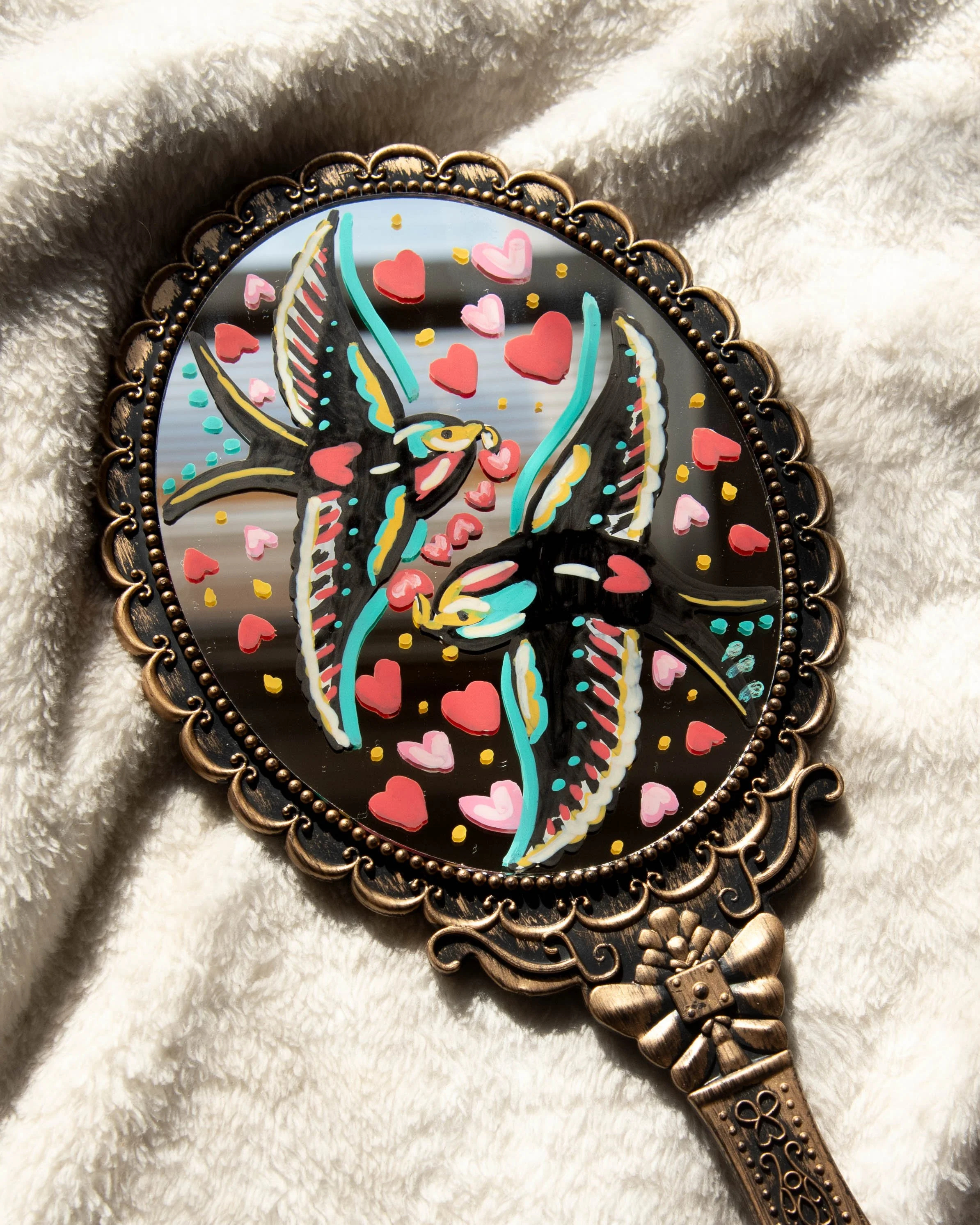 Decorative hand mirror with a colorful, vibrant design of two birds and floating hearts on the reflective surface.