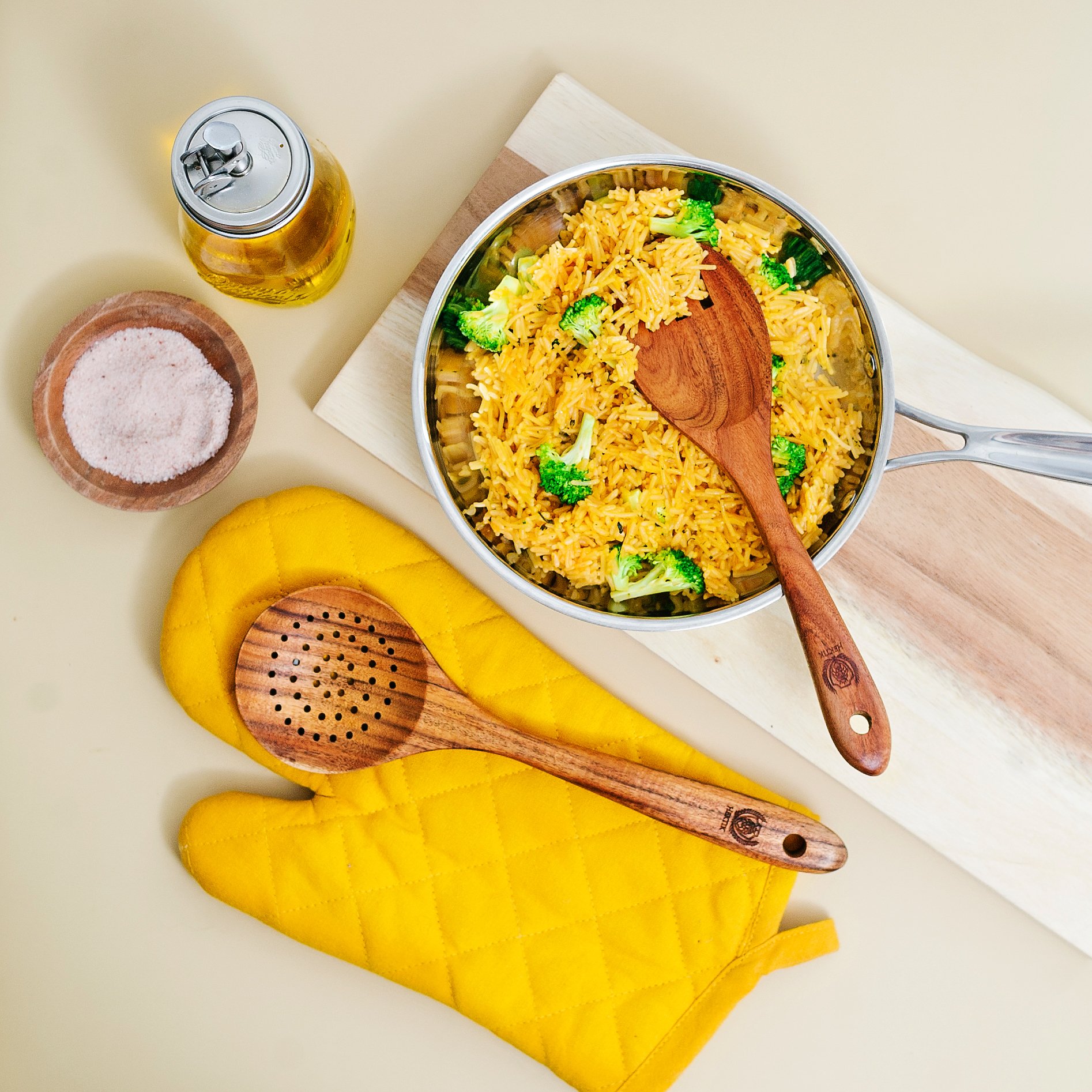 A pot of yellow rice with broccoli, a wooden spoon inside, on a white cutting board. Nearby are yellow oven mitts, a small wooden bowl of salt, a glass bottle of oil, and a long wooden spoon.