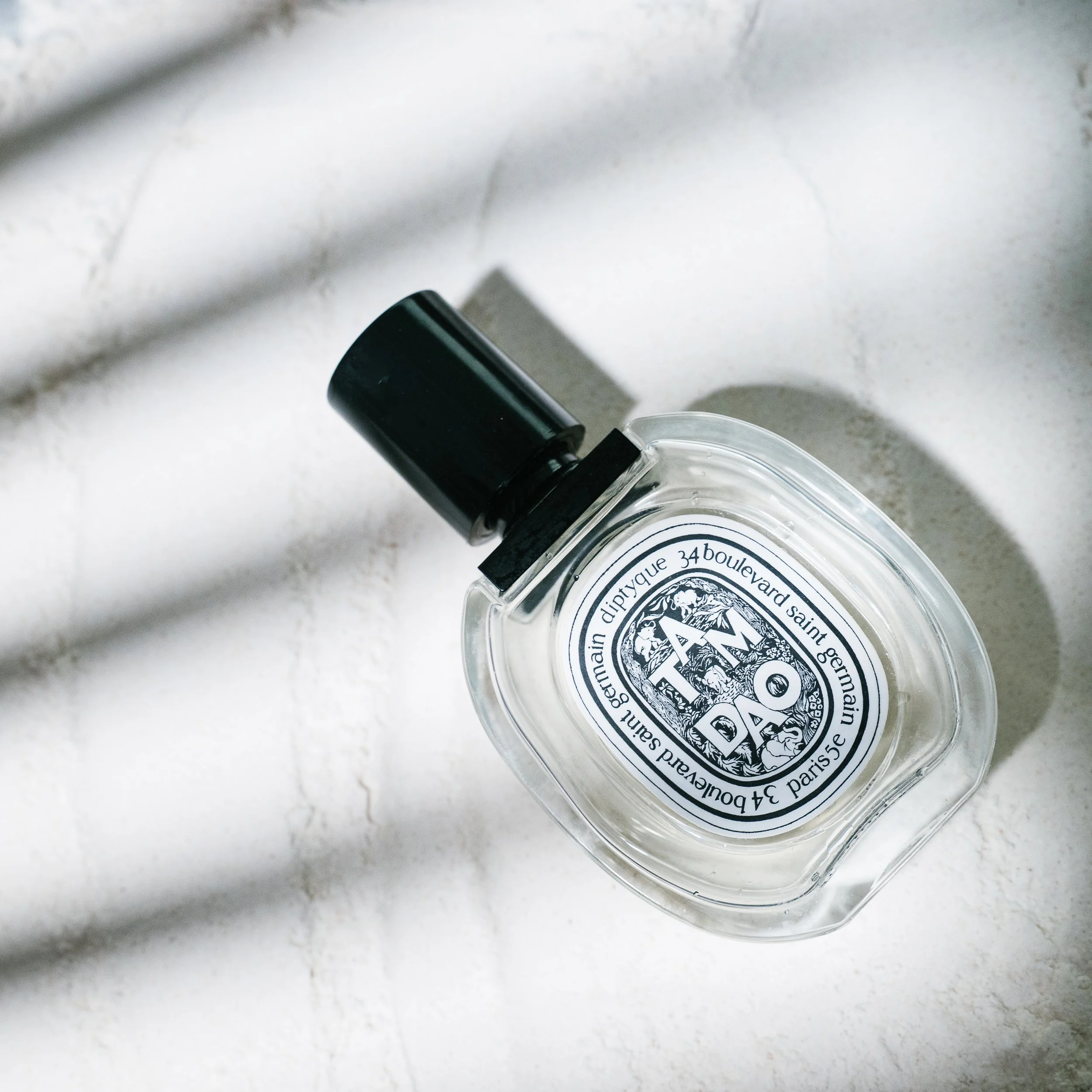 A bottle of Diptyque perfume lying on a textured white surface with shadows from nearby objects.