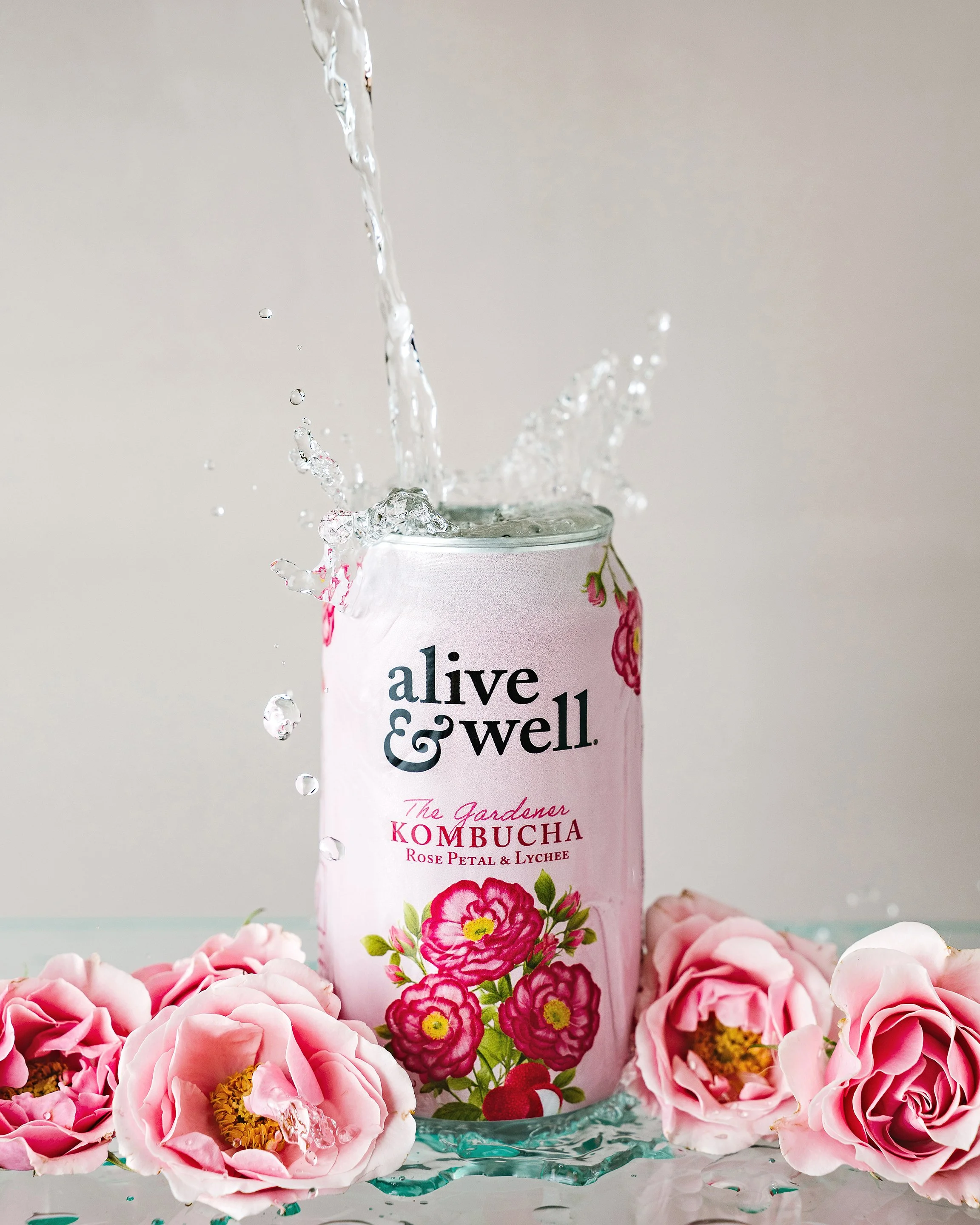 A can of live well kombucha with pink roses and petals surrounding it, as water overflows from the can with some splashing.