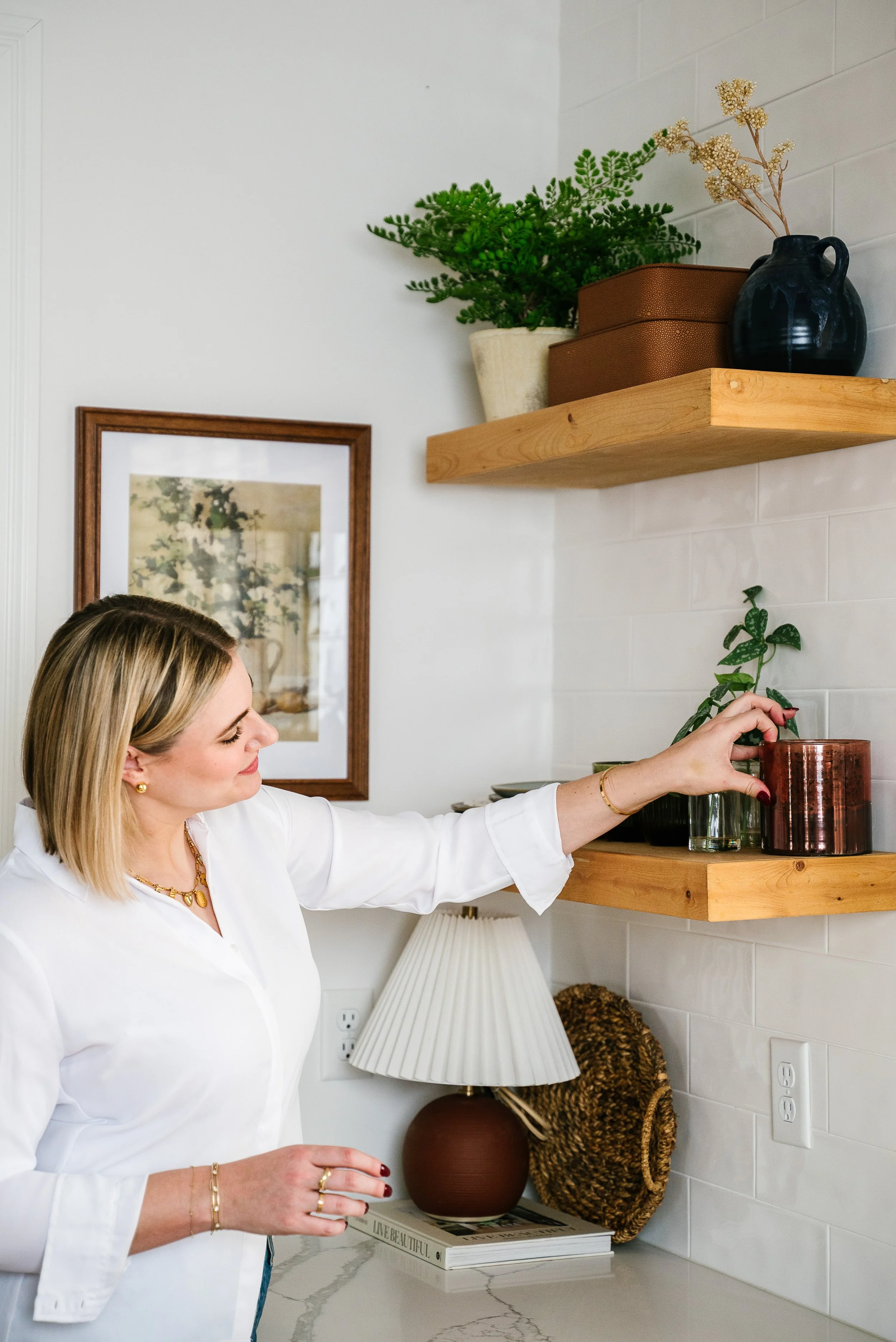 A woman with blonde hair, wearing a white blouse, arranging plants on wooden shelves in a cozy, decorated space with framed art, a table lamp, and various decorative items.