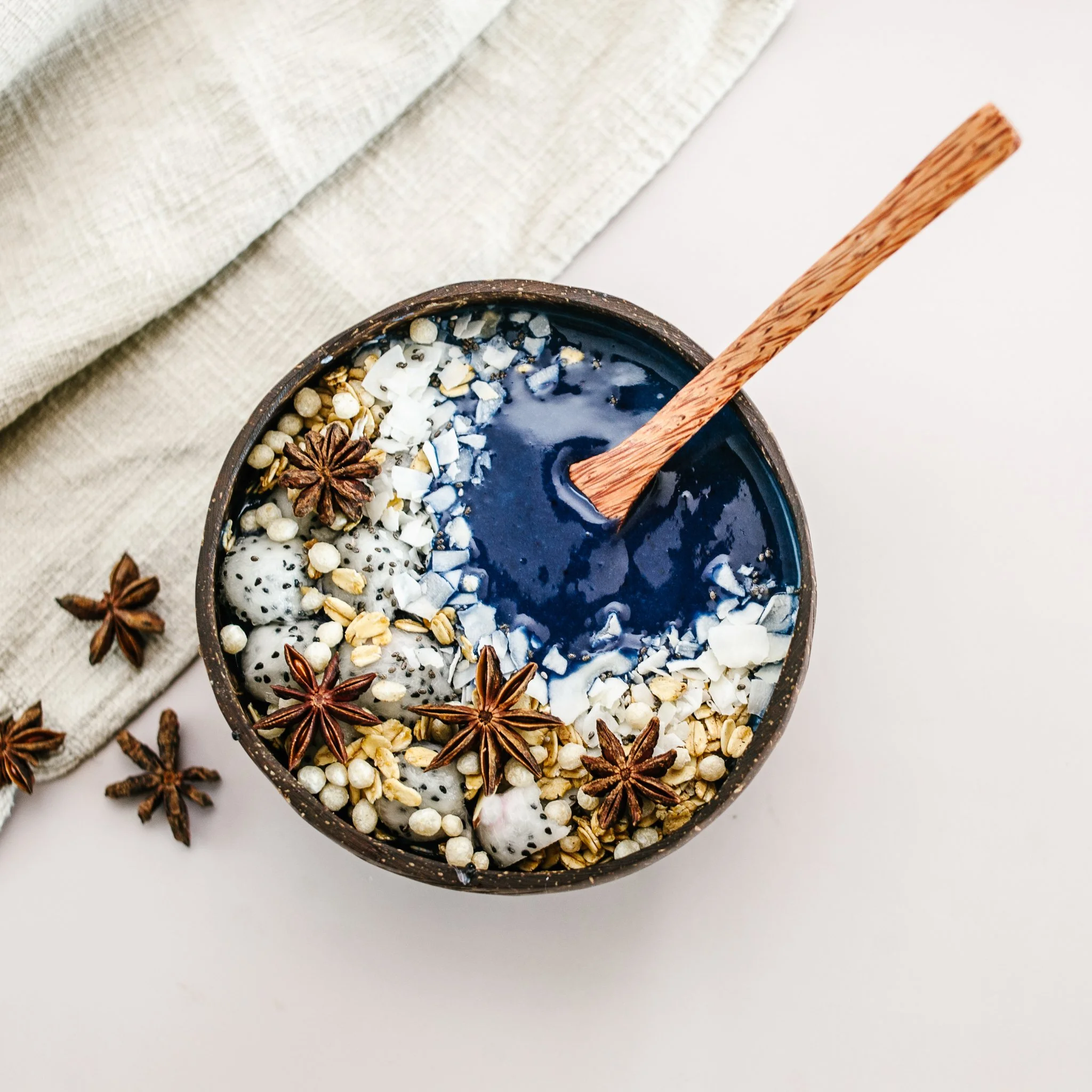 A bowl of smoothie topped with star anise, coconut flakes, chia seeds, rolled oats, and dragon fruit, with a wooden spoon inside.