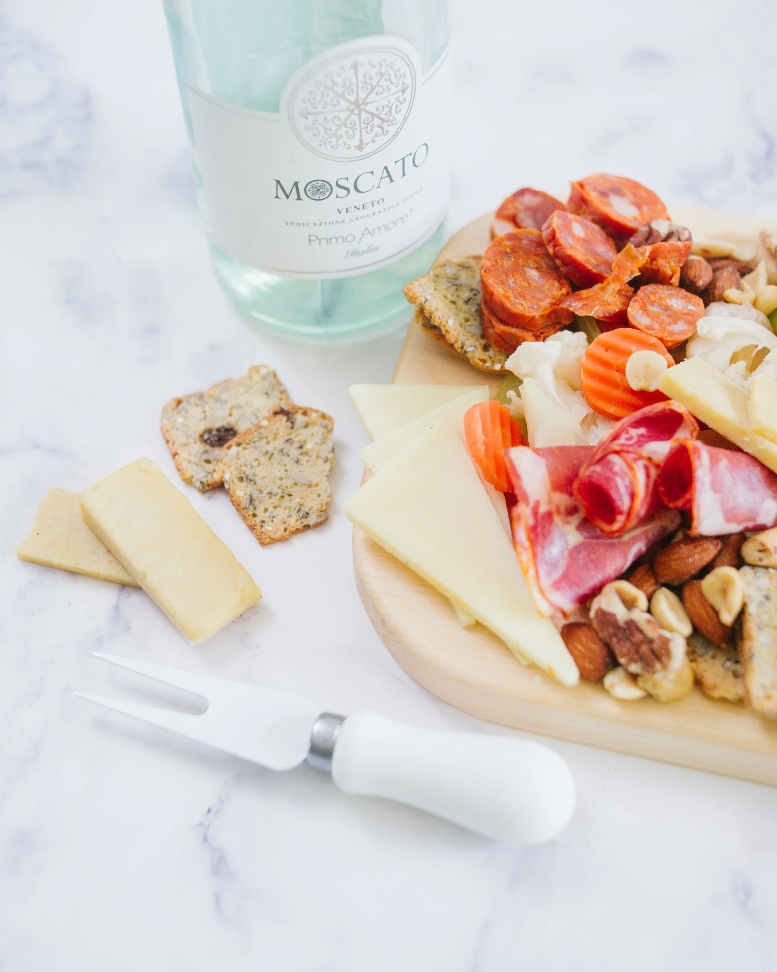Assorted cheeses, cured meats, nuts, and crackers on a wooden serving board with a cheese fork nearby. A glass of sparkling water in the background.