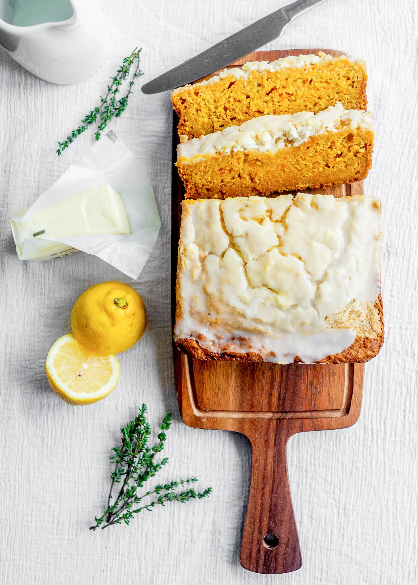 Lemon pound cake slices on a wooden serving board with lemon halves, butter, a sprig of thyme, and a knife on a white textured surface.