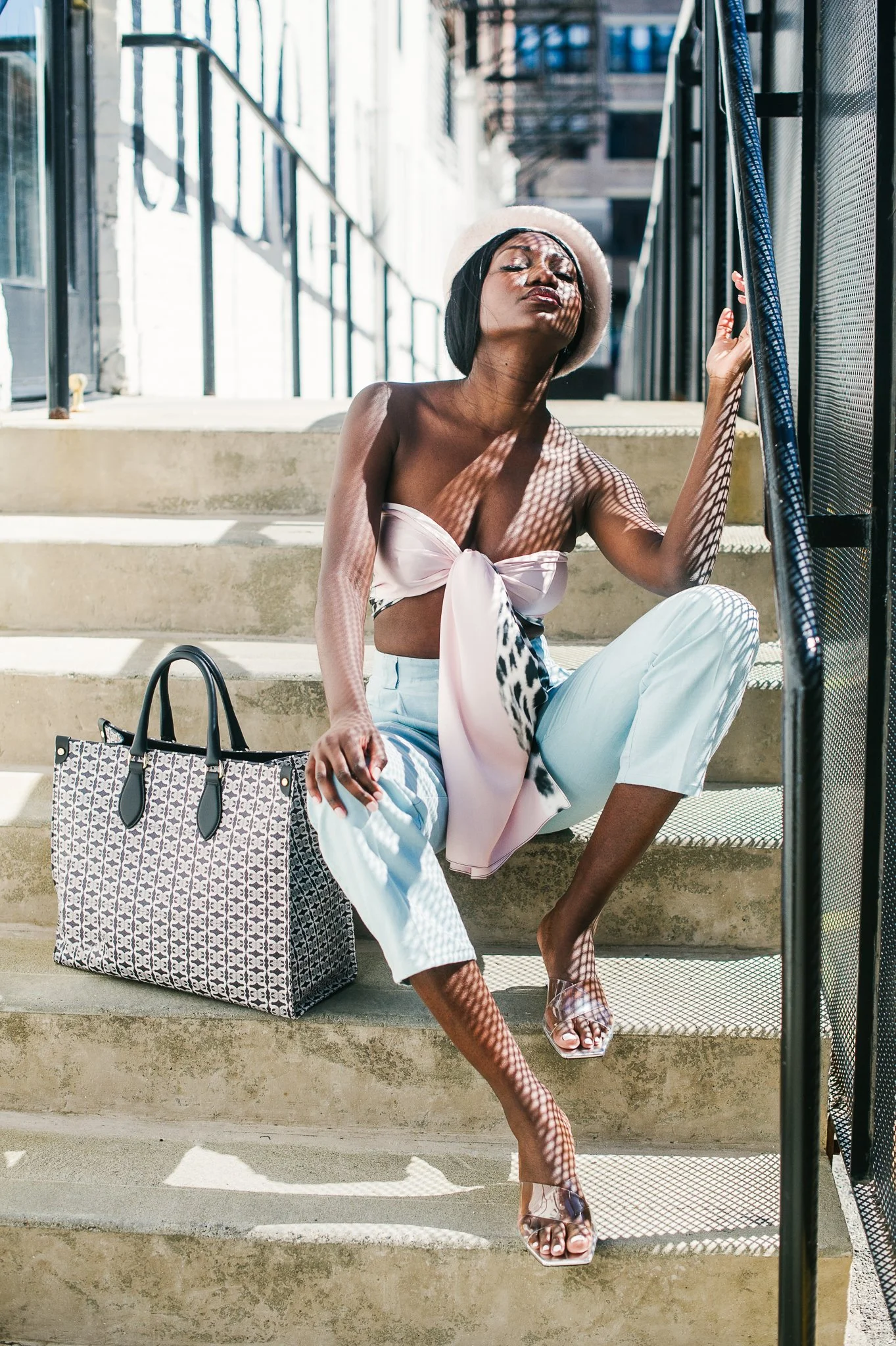 A woman sitting on outdoor concrete stairs in sunlight, wearing a light pink bandeau top with a bow, white pants, clear high heels, and a beige hat, with a patterned tote bag beside her, sun casting shadows through a metal railing.