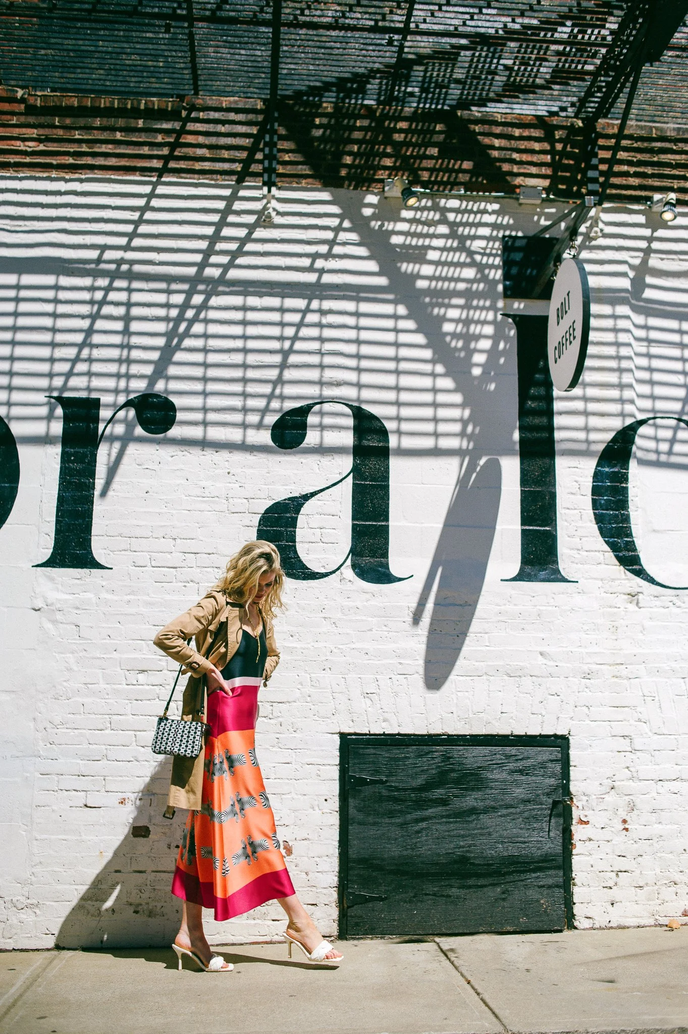 A woman with blonde hair wearing a colorful dress, tan coat, white high heels, and carrying a handbag, stands against a white brick wall with large painted lettering. Shadows and the shadow of a fire escape are cast onto the wall, adding visual inter
