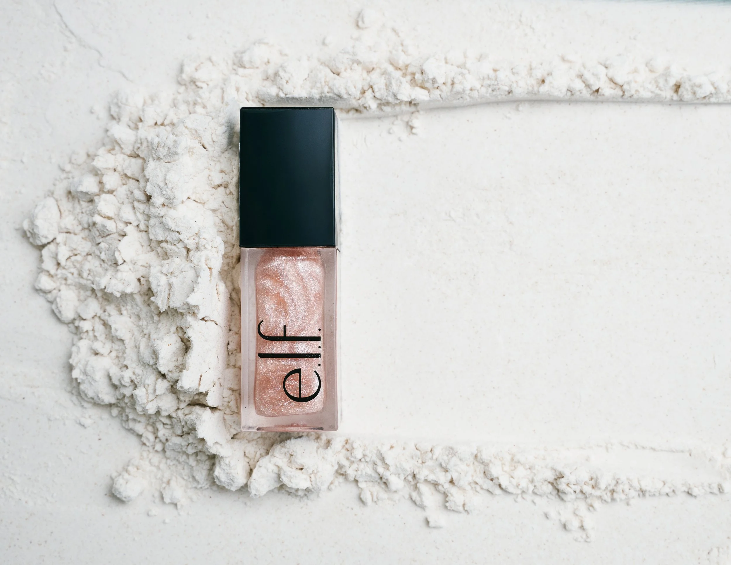 A bottle of e.l.f. cosmetics nail polish with a black cap, lying on a white surface surrounded by white powder.