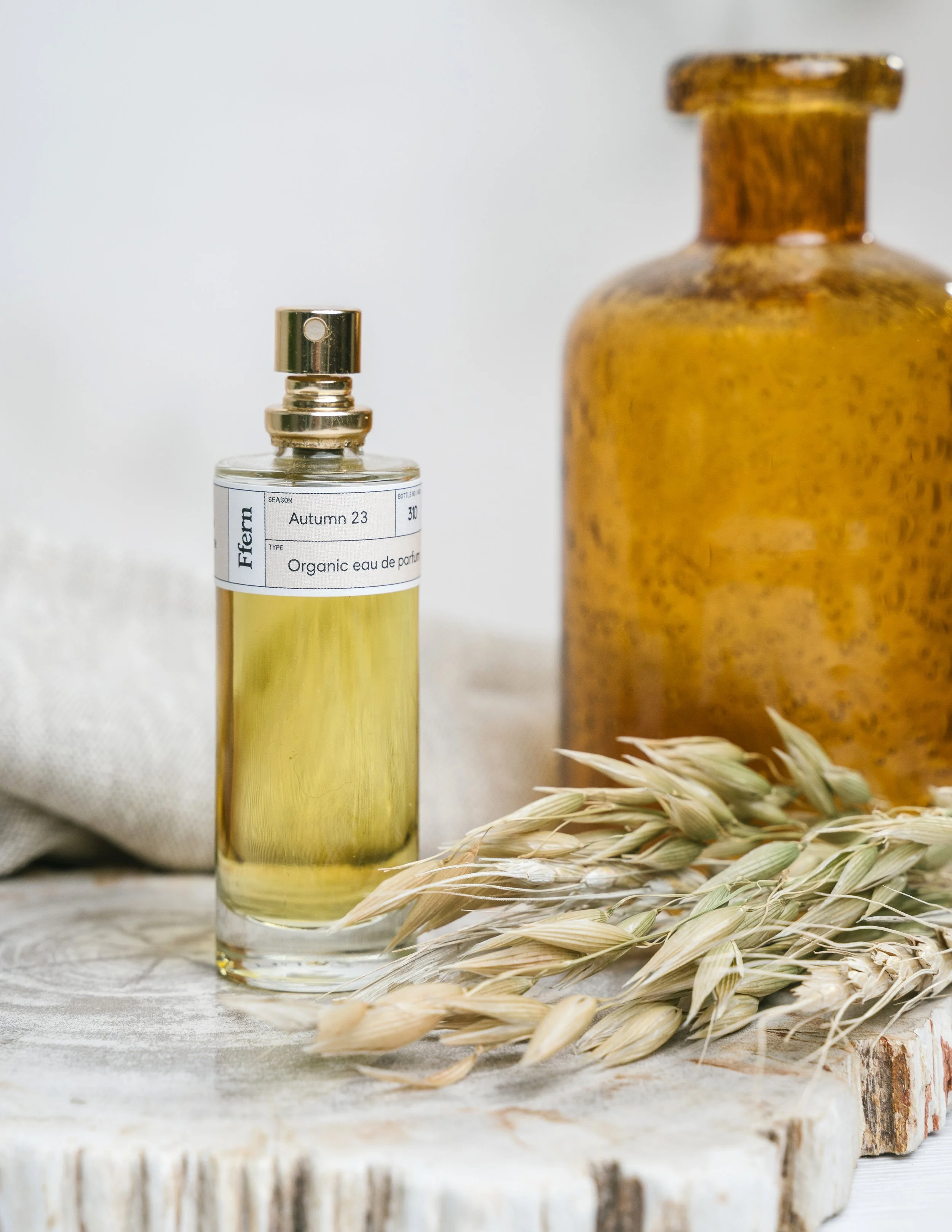 A glass perfume bottle with a metallic spray top and a label that reads 'Autumn 23, Organic eau de parfum', a large amber-colored bottle, and dried wheat stalks on a wooden surface.
