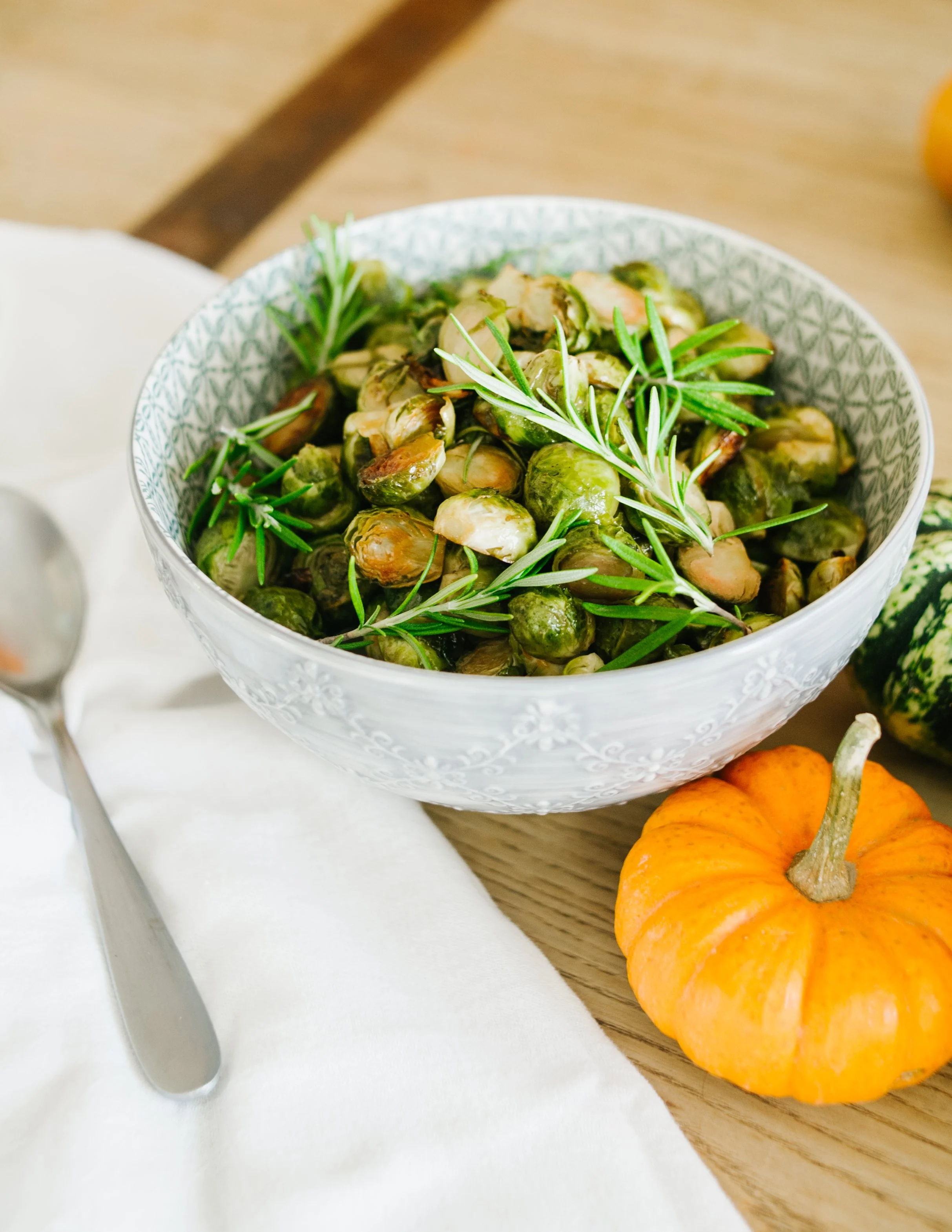 A decorative glass bowl filled with roasted Brussels sprouts garnished with fresh rosemary sprigs, placed on a wooden table with a pumpkin nearby.