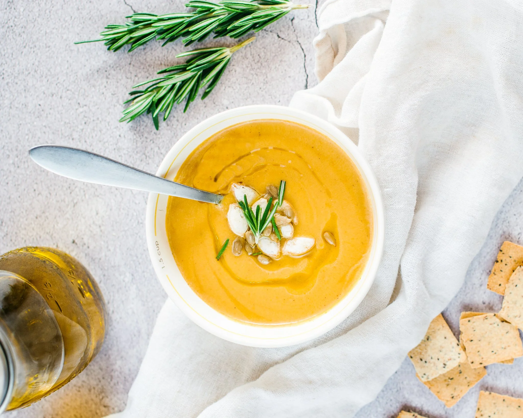 A bowl of creamy orange soup garnished with herbs, garlic slices, and sunflower seeds, with a spoon inside, on a white cloth next to croutons and a sprig of rosemary