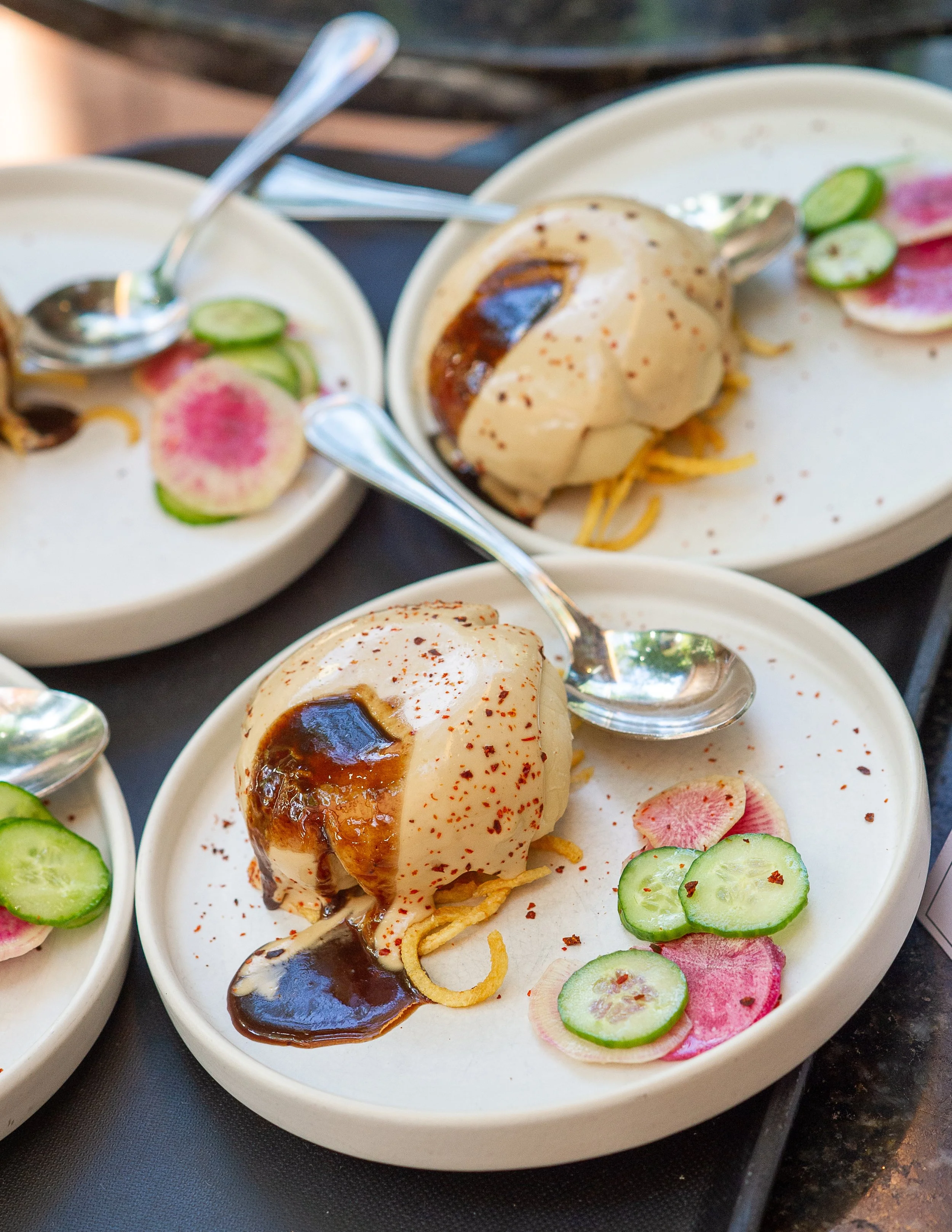 Three white plates with dessert featuring vanilla ice cream topped with chocolate sauce, served with sliced cucumbers and pickled radishes.