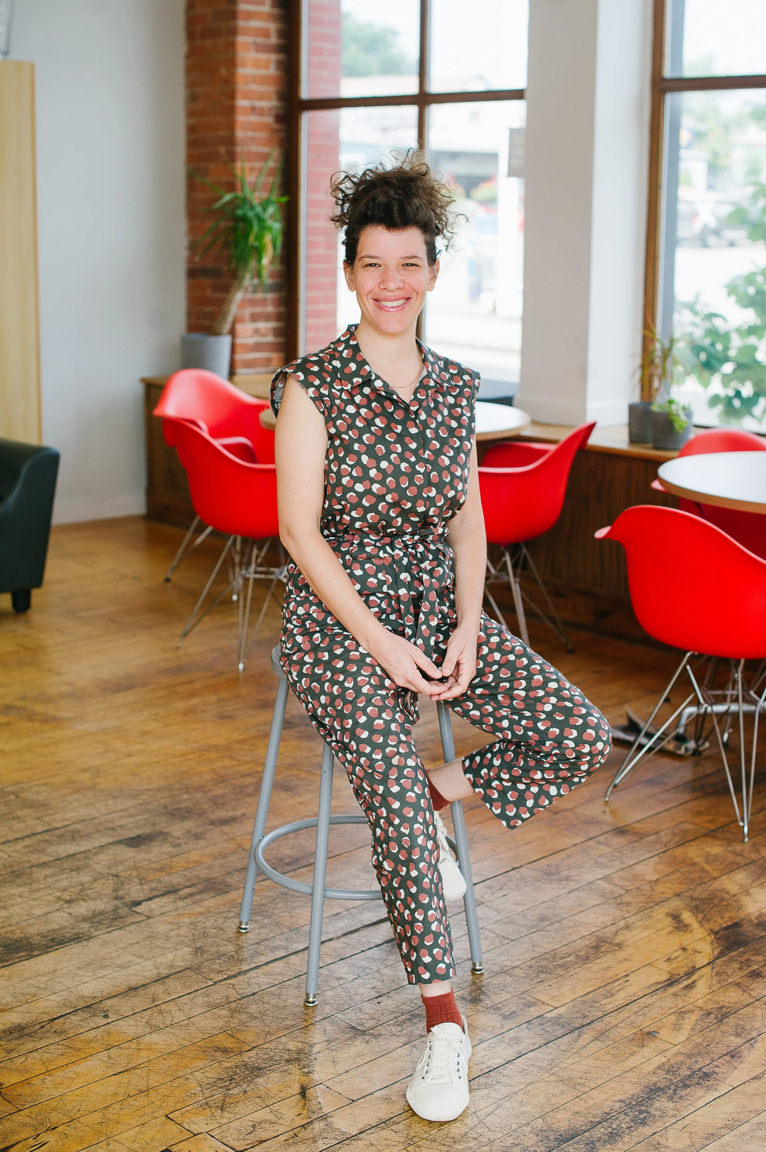 Woman sitting on a stool in a brightly lit modern cafe with red chairs and large windows.
