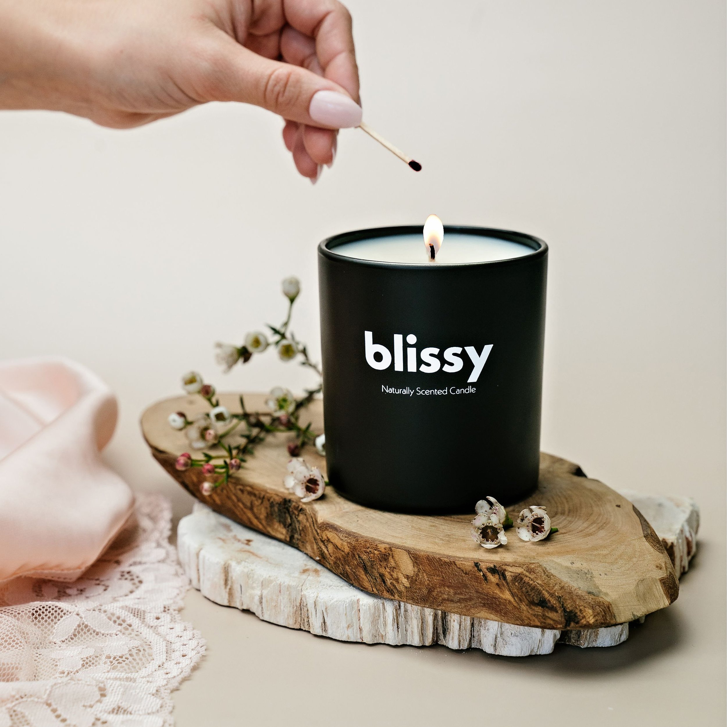 A hand lighting a black scented candle labeled "blissy" with a matchstick. The candle is on a wooden slab decorated with small white flowers, on a soft pink lace cloth.