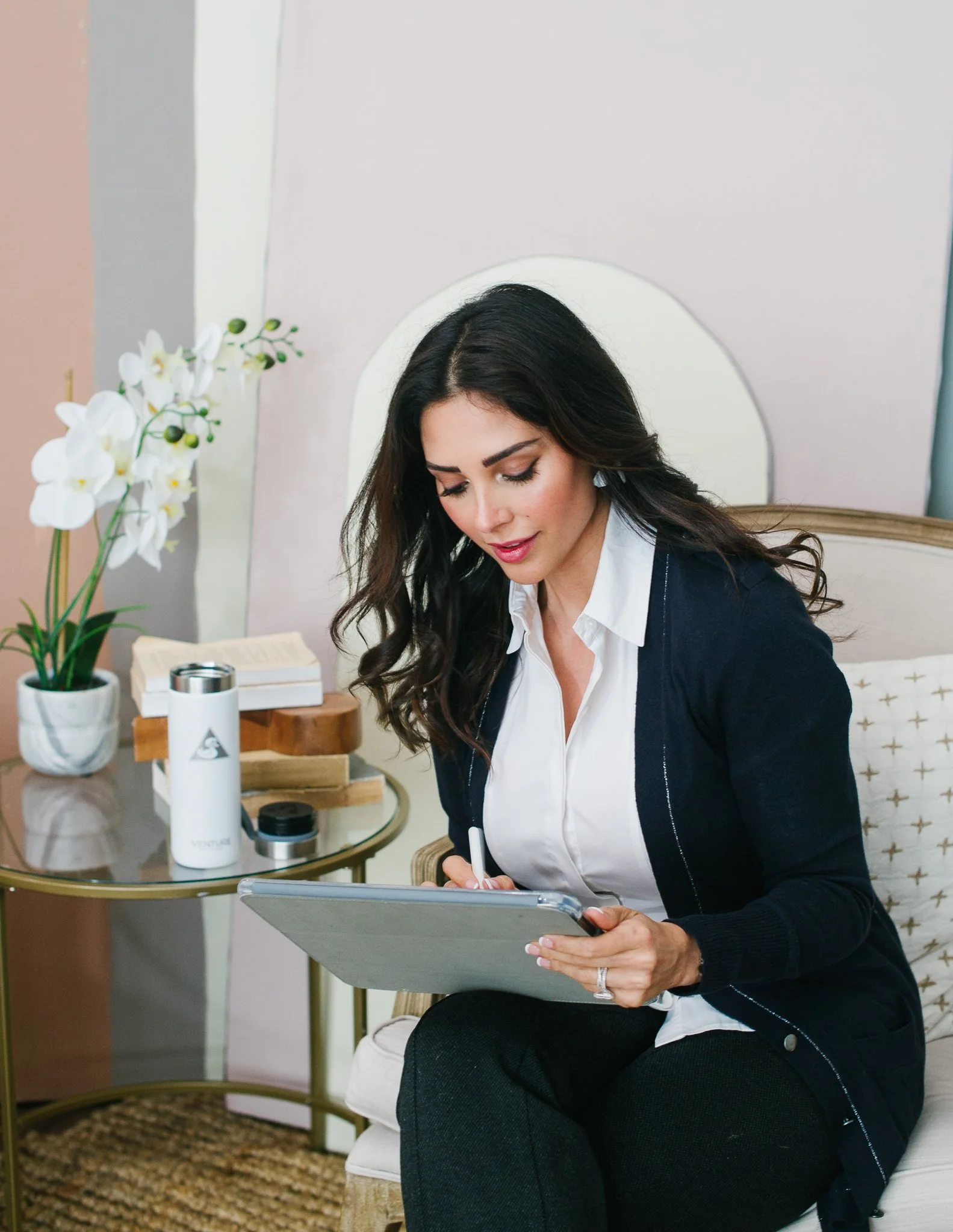 A woman sitting on a cream-colored chair, writing on a clipboard. There is a small gold side table beside her with a potted orchid, a tea bottle, and some books.