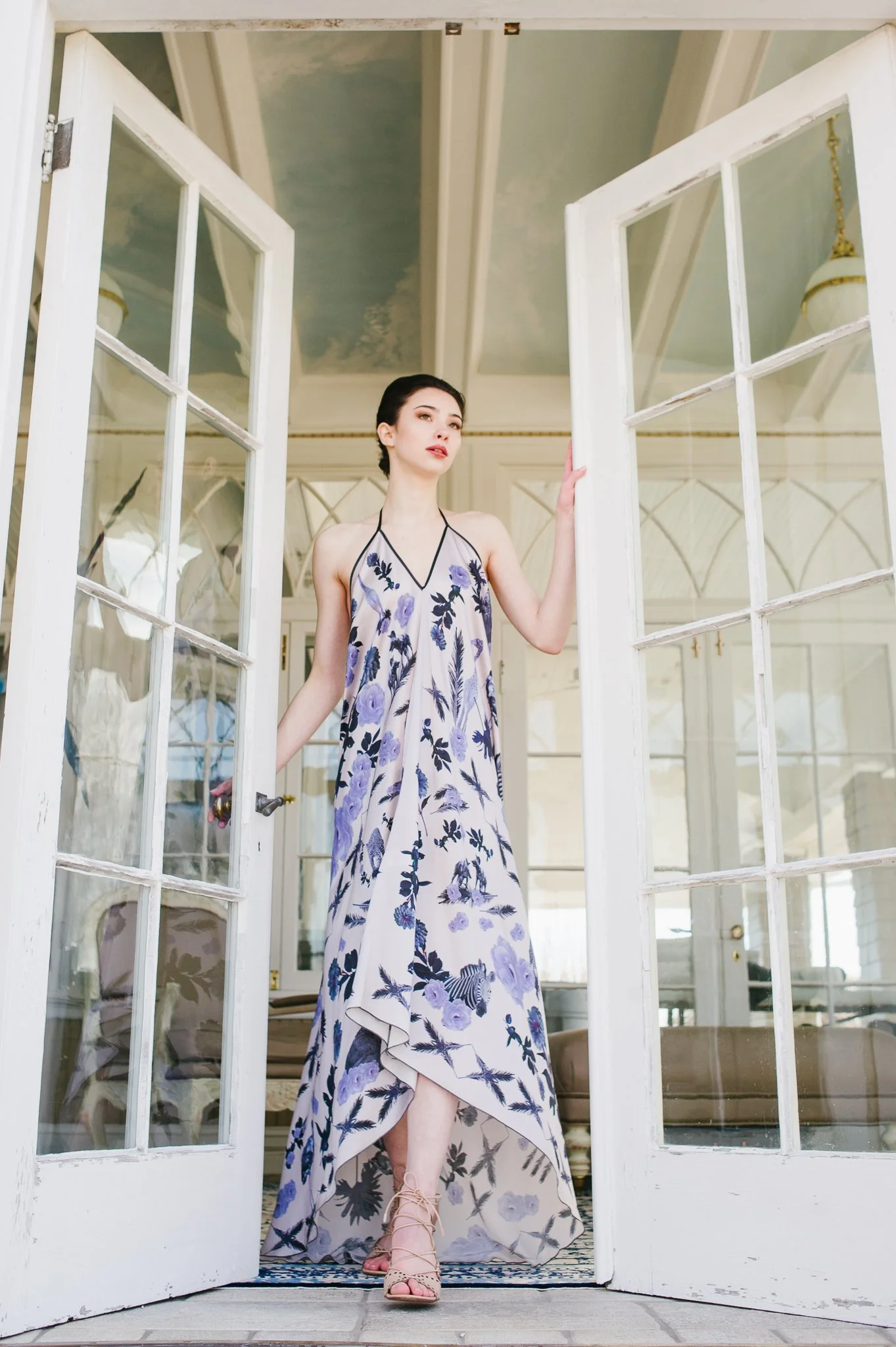 A woman in a long, floral dress standing in a doorway with white glass-paneled doors, inside a bright and elegant room with decorative ceiling and chandeliers.