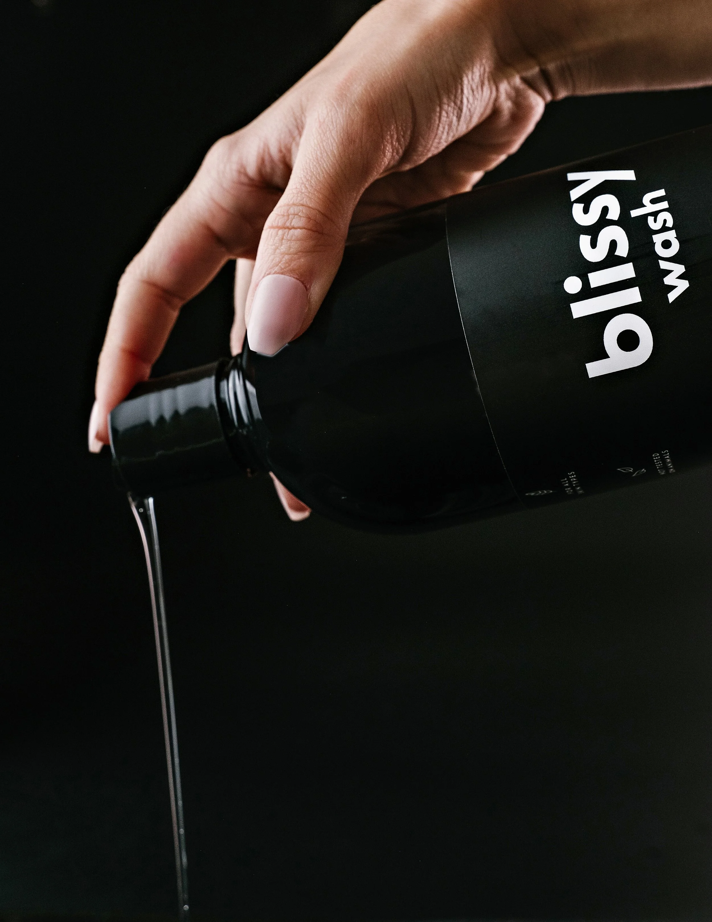 Close-up of a hand with manicured nails pouring liquid from a black bottle labeled 'blissy wash' onto a black surface.