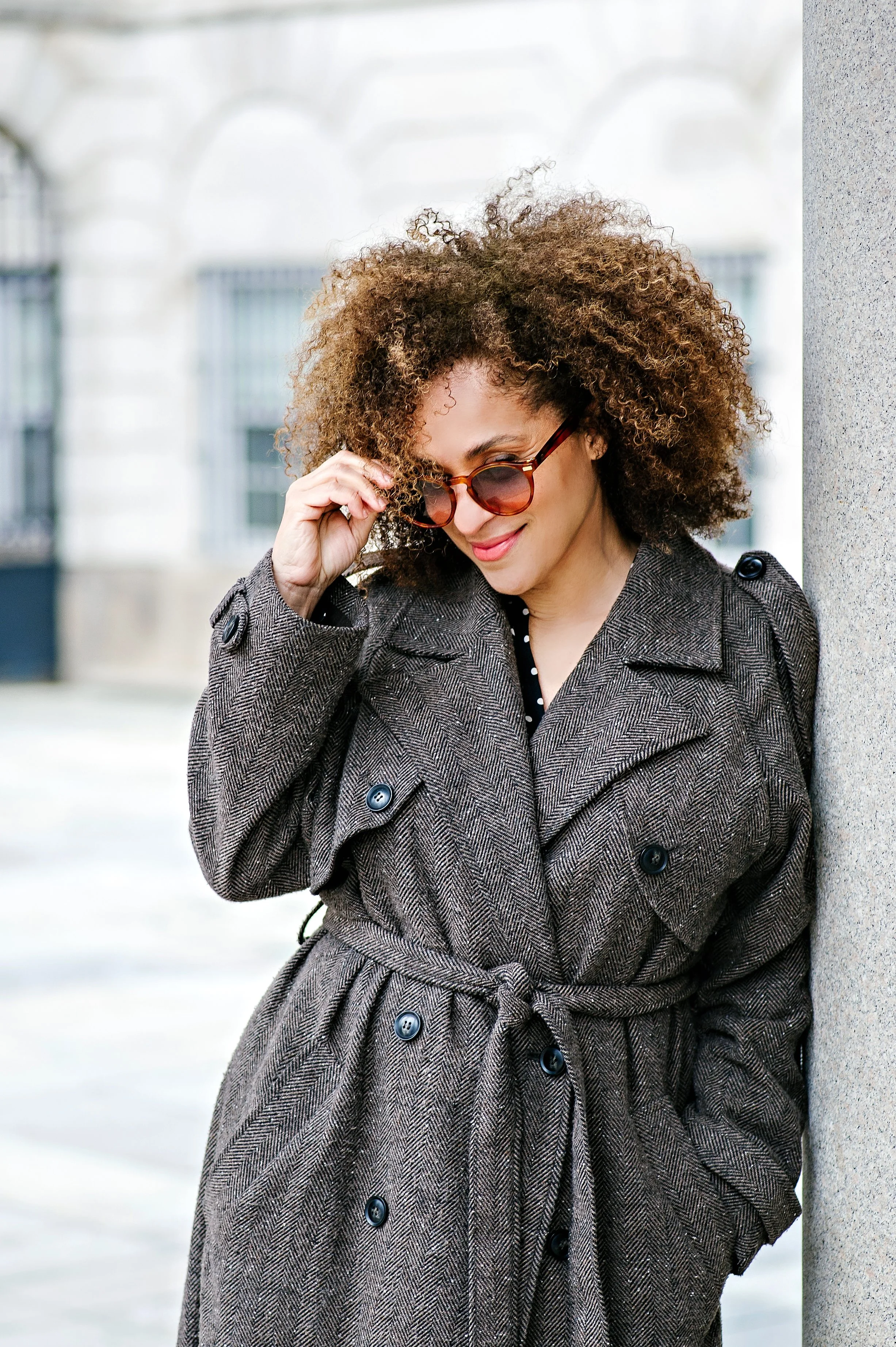 A woman with curly hair wearing sunglasses and a brown trench coat standing against a stone wall on a city street.