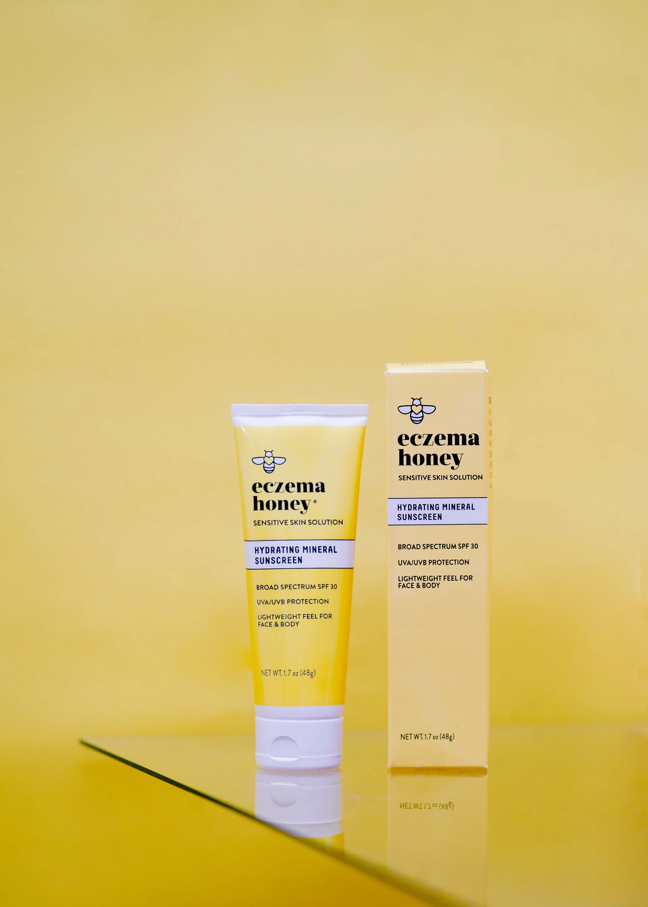 A tube of eczema honey hydrating mineral sunscreen with broad spectrum SPF 30 and UVA/UVB protection, placed next to its yellow box packaging on a yellow surface with a yellow background.
