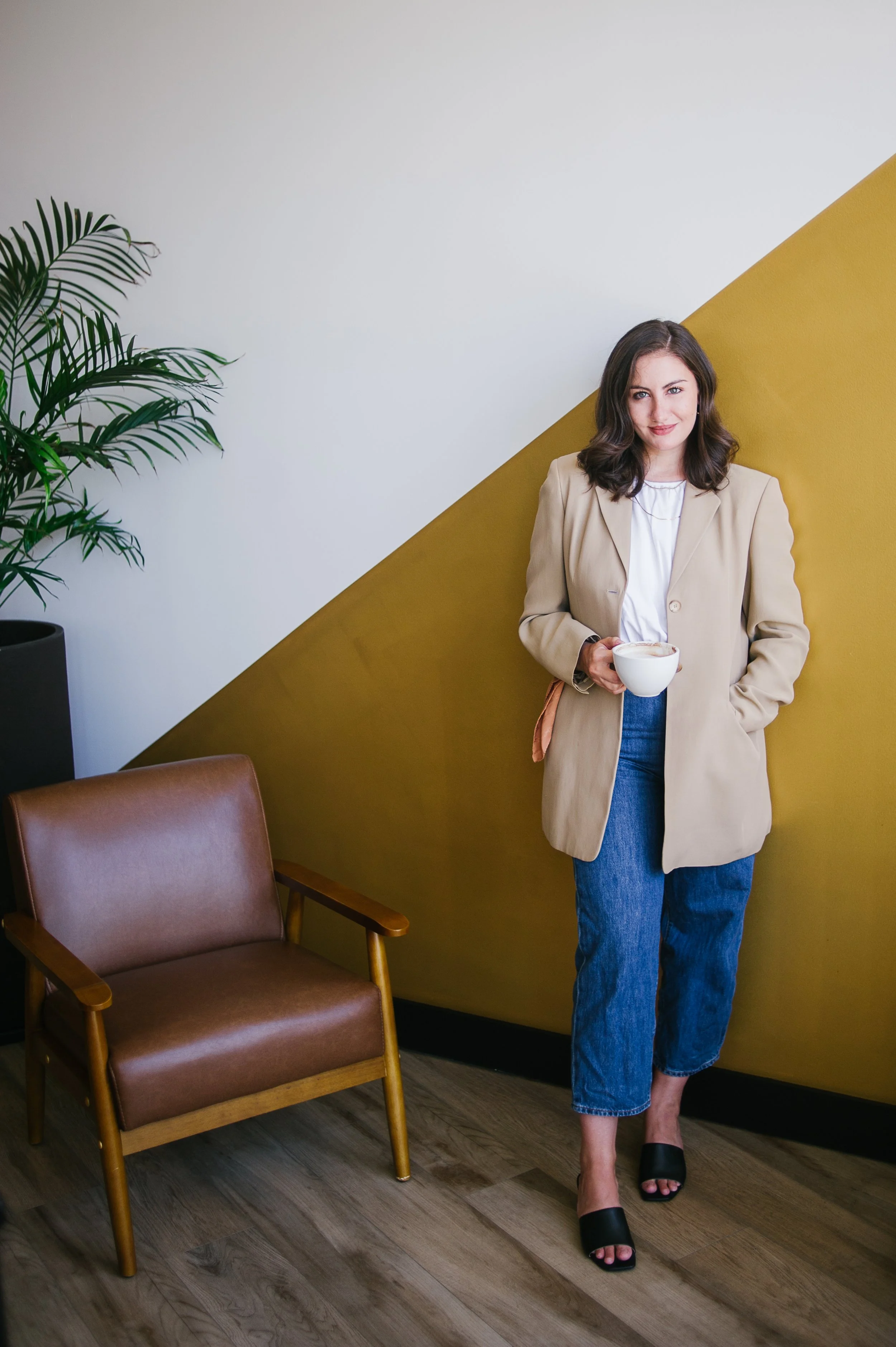 Branding photography featuring a woman with dark hair, wearing a beige blazer, white shirt, and blue jeans, standing in a room with a brown leather chair, a potted plant, and a yellow wall, holding a white mug. 