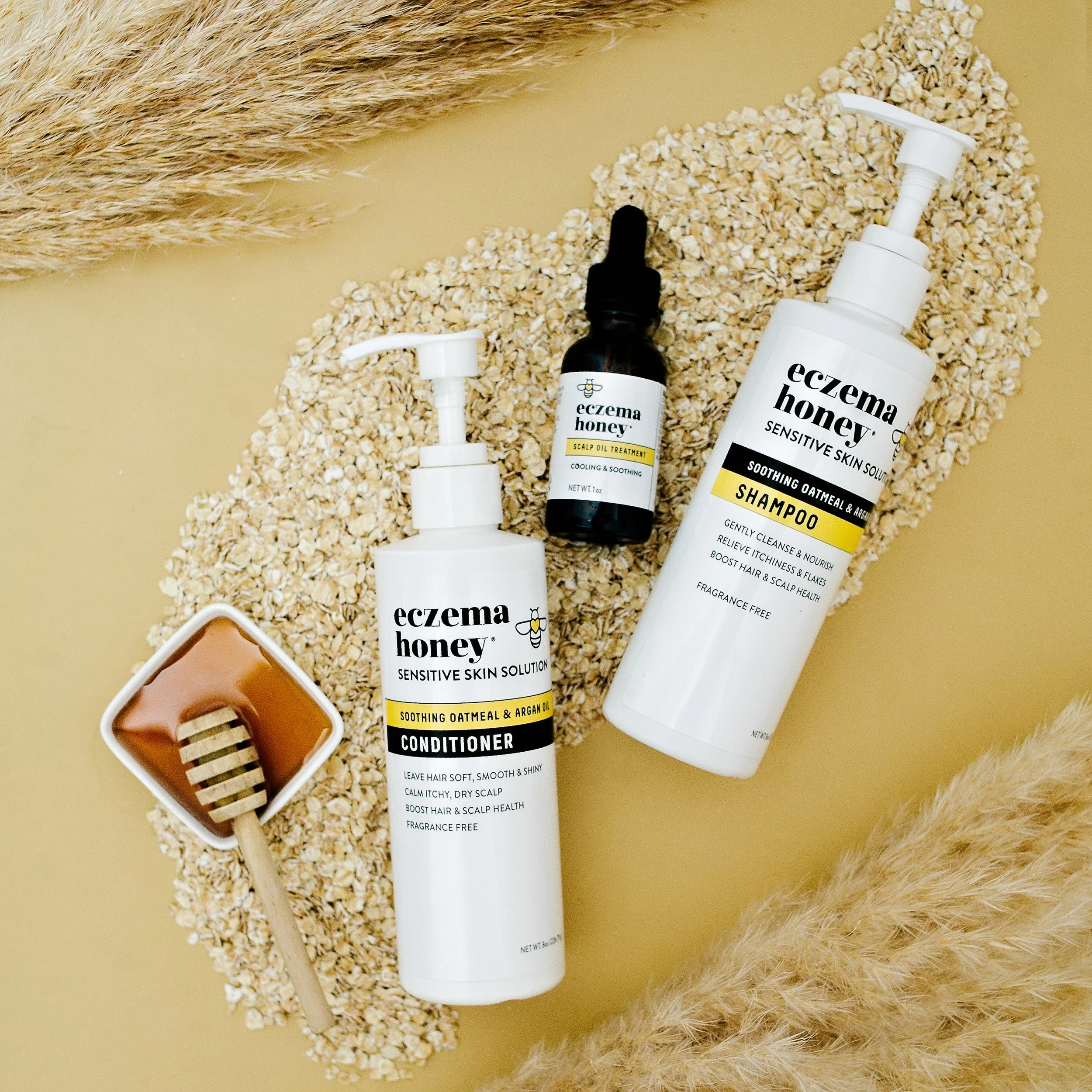 Eczema Honey skincare products including a conditioner, shampoo, and scalp oil treatment, arranged on a beige surface with dried wheat and oats for a natural aesthetic.